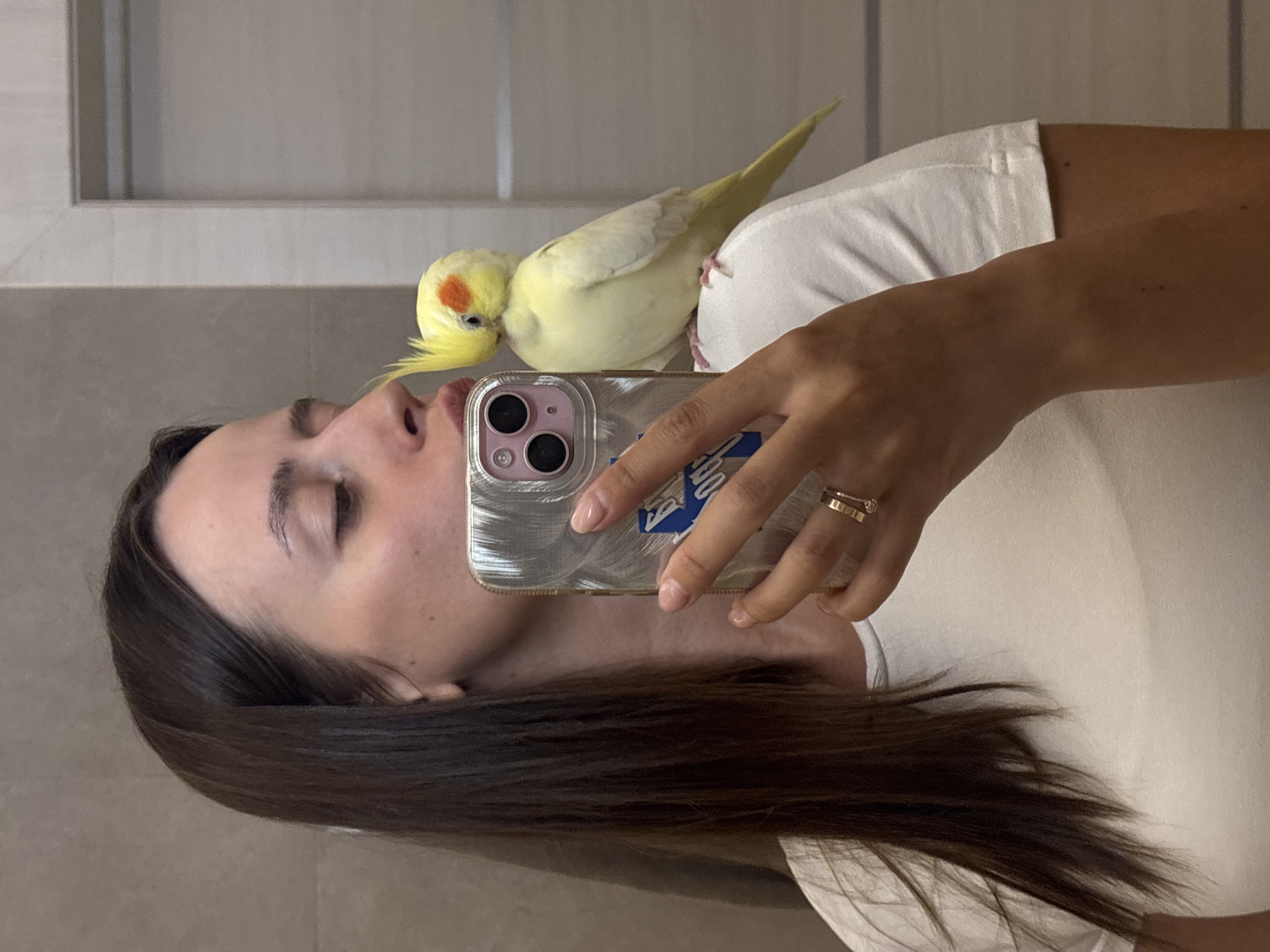 The image shows a woman taking a selfie in a bathroom. She is wearing a white t-shirt and has long, dark brown hair. A light yellow cockatiel with an orange cheek patch is perched on her shoulder, close to her face. The woman is leaning in, as if to kiss the bird, and is holding her phone up to take the picture. The phone has a silver case and a blue sticker. She is wearing a ring on her right hand. The background appears to be a tiled wall, suggesting a bathroom or similar room. The image does not contain any information that would indicate its location in Krasnoyarsk, Russia, or any other specific city.