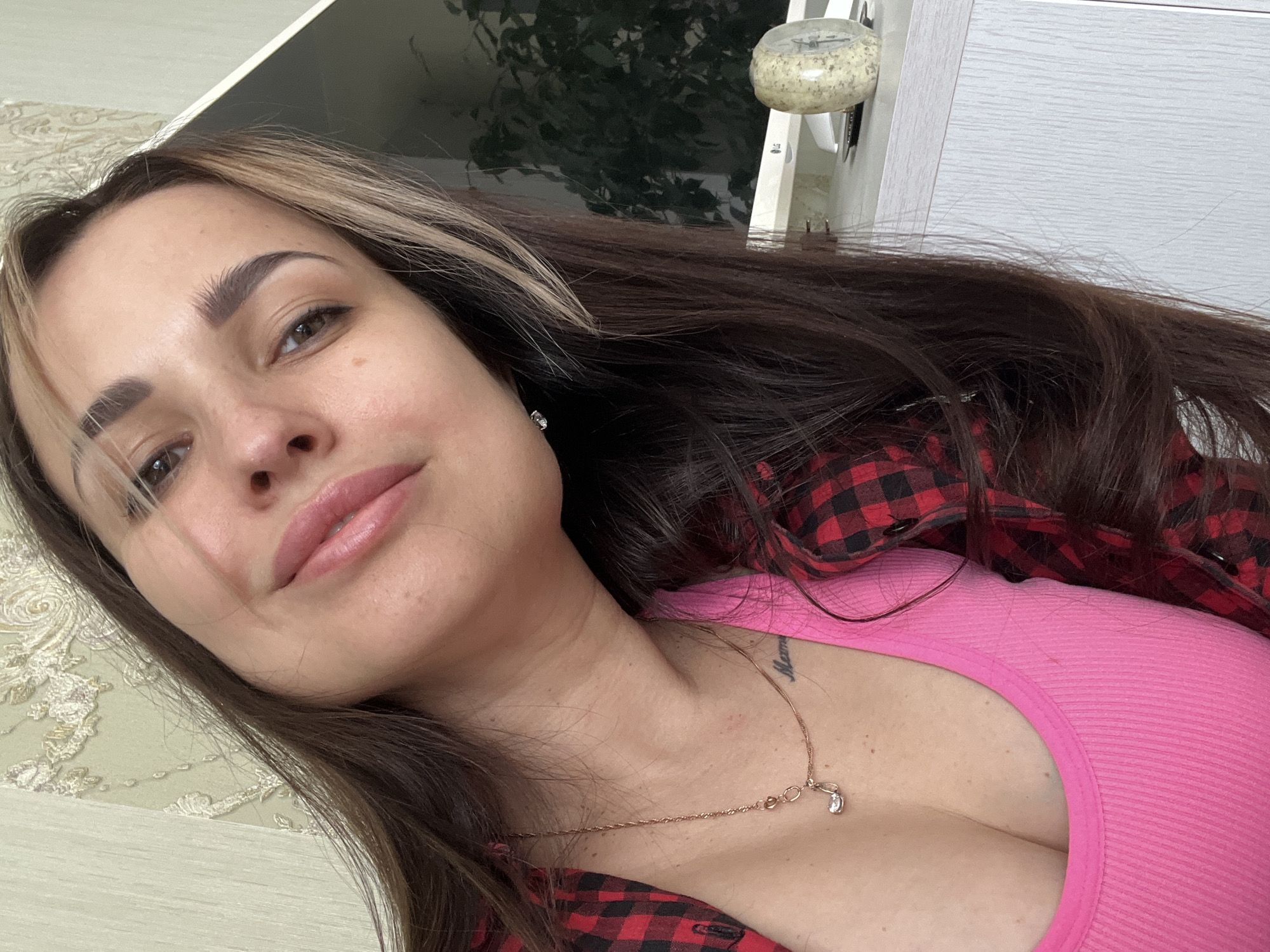 A woman with long, dark hair, featuring lighter streaks, poses for a photograph indoors. She is looking directly at the camera with a relaxed, slight smile. Her eyebrows are well-defined, and she has light makeup on her eyes and lips. She is wearing a bright pink, ribbed tank top, underneath an open red and black plaid button-up shirt. Small stud earrings are visible, and she wears a delicate gold-toned chain necklace with a small, clear stone pendant. Faint dark text, possibly a tattoo, appears on her left collarbone, partially obscured by the necklace, which seems to spell "Mama".
The background suggests a domestic setting, with a dark, reflective surface (possibly a table) to her right, reflecting a blurry green shape that resembles a plant. A white cabinet or wall unit is also visible, with a small, round, mottled object placed on top of it. The wall to her left features an intricate, light-colored pattern. The image is taken at an angle, giving a dynamic perspective to the scene. The lighting suggests it is daytime.