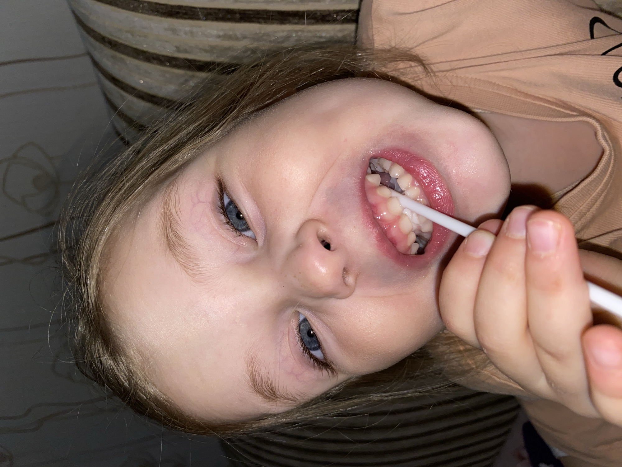 The image provides an overhead, close-up view of a young child, likely a girl, engaged in dental hygiene. The child has light brown or blonde hair and striking blue eyes, looking directly at the camera with a focused and somewhat serious expression. Her mouth is open, revealing her upper and lower primary teeth. A small white brush or swab is positioned against her teeth, held by what appears to be her own small hand, indicating she is actively participating in or performing the brushing herself. She is dressed in a light peach or orange-colored top.
The setting is indoors, with a striped upholstered surface visible behind her head, possibly a sofa or chair, and a darker, subtly textured background to the left. The lighting is soft and even, typical of an indoor environment. The scene captures a routine personal care moment. While the geographic context provided is Votkinsk, Russia, no specific visual cues within the image itself identify this location. There is no visible text in the image.