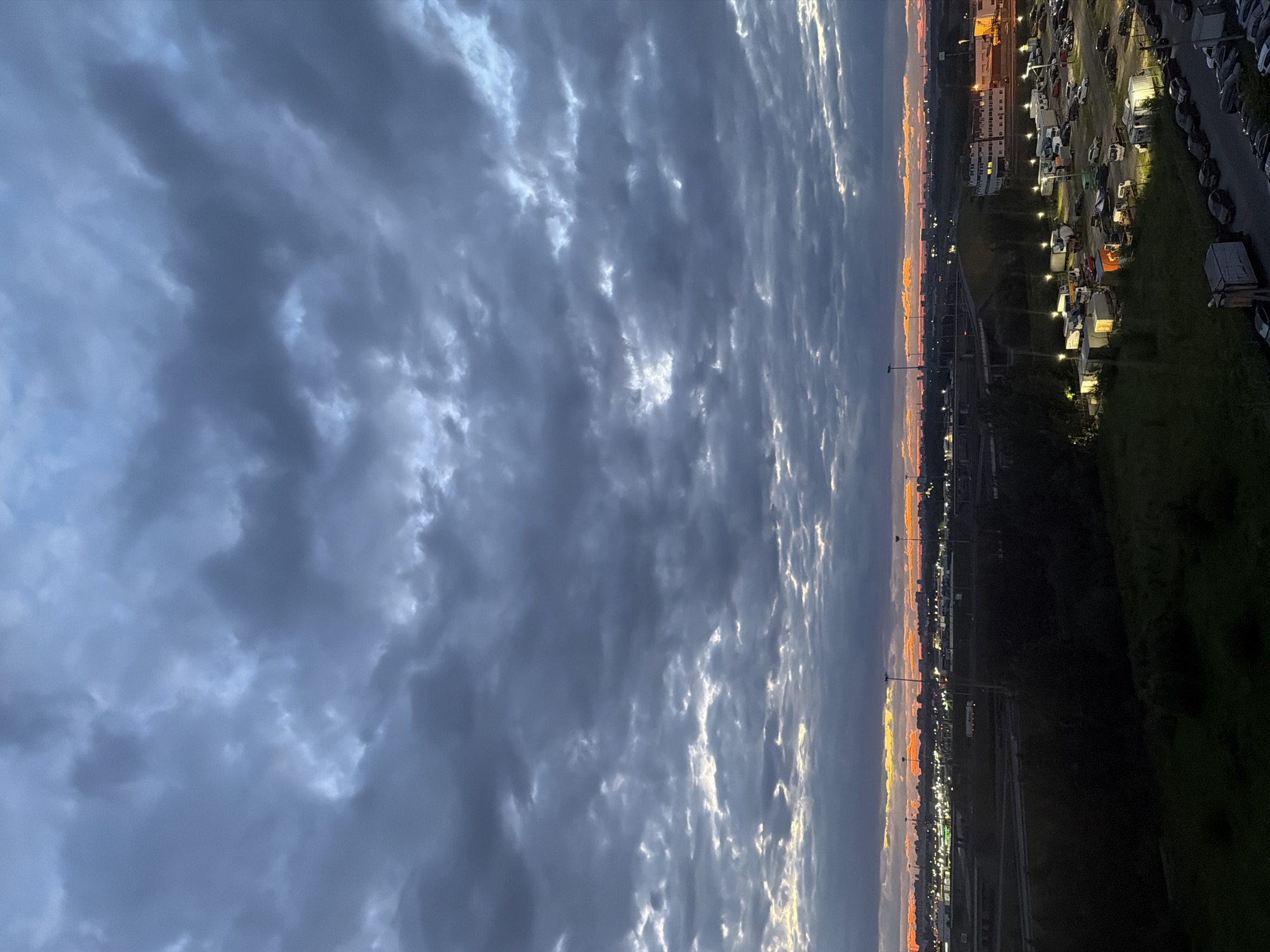 The image depicts a dusky sky with dramatic, dark clouds in the upper portion, broken by streaks of light orange and yellow near the horizon. Below the sky, a cityscape is visible, characterized by a dense collection of buildings, streetlights, and what appears to be a parking lot filled with numerous vehicles, including cars, trucks, and RVs. There are also some industrial-looking structures and what might be a highway or overpass in the mid-ground. The overall scene suggests an urban environment at twilight or dusk, with artificial lights illuminating parts of the landscape beneath the darkening sky. No people are visible in the image.