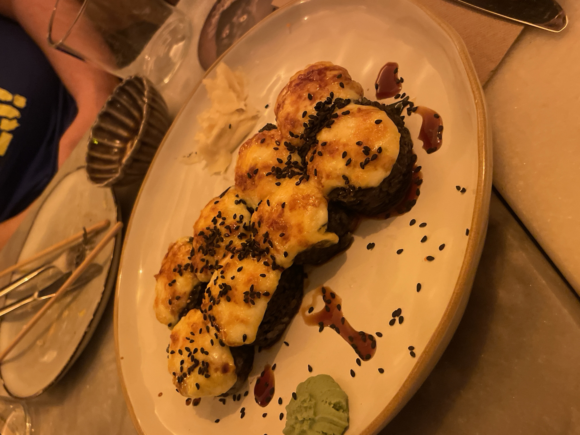 The image shows a plate of food, likely sushi, with several pieces topped with melted cheese and sprinkled with black sesame seeds. There is a small dollop of wasabi and ginger on the plate, along with some dark-colored sauce. In the background, there is a glimpse of a person wearing a blue shirt with yellow stripes, a small bowl with ridges, and used cutlery on a plate. The overall setting suggests a restaurant environment, possibly in Barcelona, Spain. The lighting is warm, indicating an indoor setting.