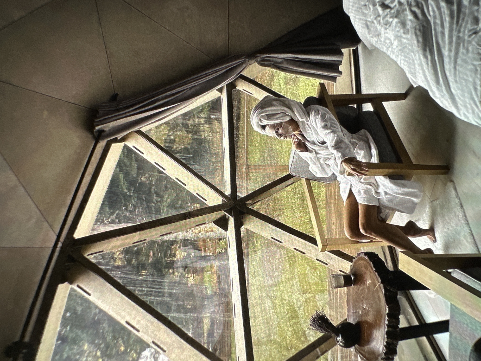 A woman wearing a white robe and a towel wrapped around her head sits in a wooden chair inside a geodesic dome structure with triangular window panes revealing an outdoor view with greenery. A small round table with objects on it sits to the right of the chair. The inside of the dome is partially obscured by a gray curtain at the top. A bed is located at the top right corner of the photo. The image appears to be taken with a camera tilted on its side. The location is not specified in the prompt, and it is unknown if it is in Yerevan, Armenia.