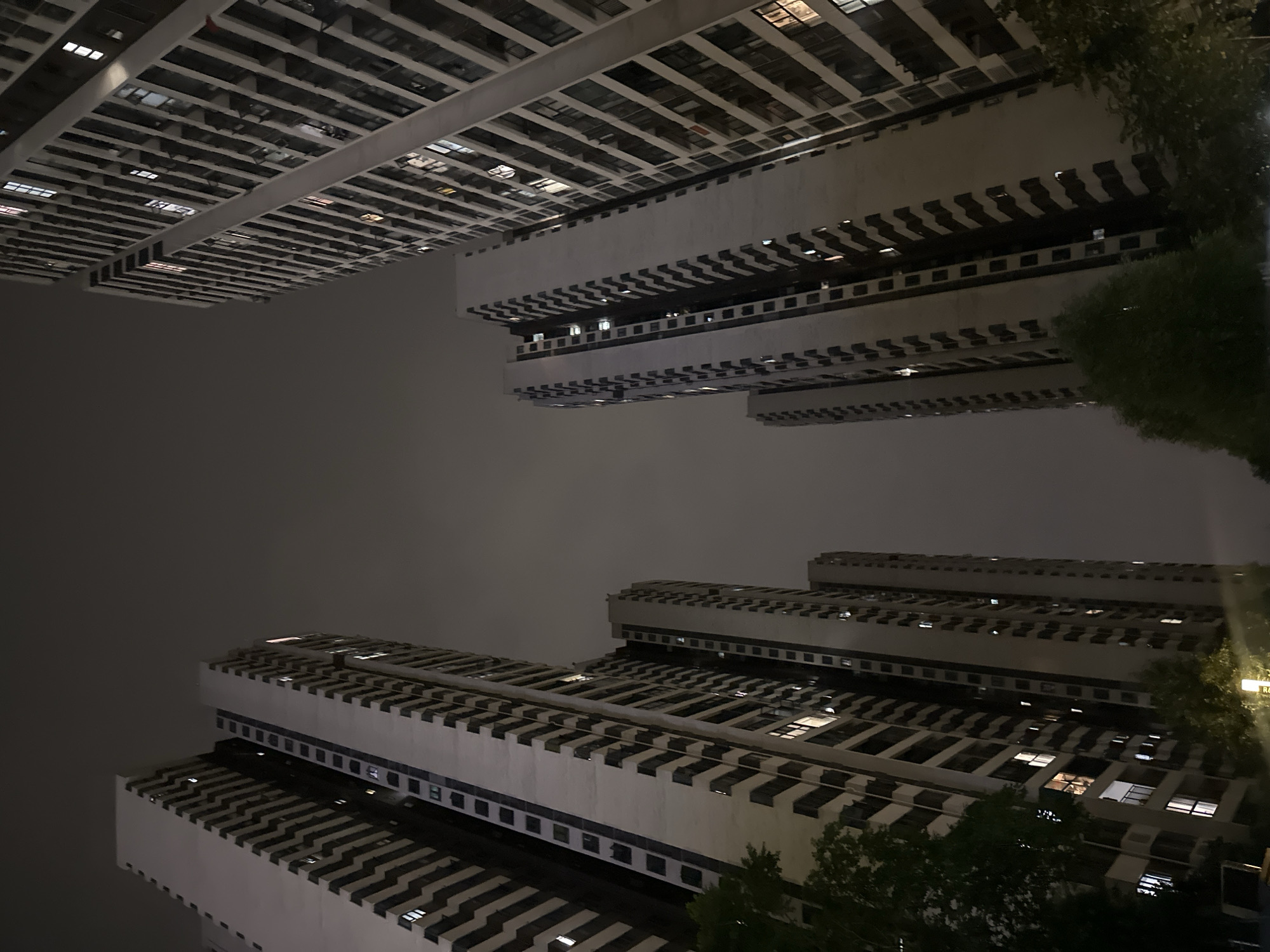 The image captures a night scene in Chongqing High-tech Zone, China. The photograph looks up at high-rise buildings, arranged in two groups that appear to mirror each other. The buildings are characterized by a grid-like architectural pattern, with alternating stripes of light and dark that likely correspond to windows and structural elements. Some windows are illuminated, indicating occupancy. The sky is overcast, creating a muted lighting environment. In the foreground, there are the tops of trees, suggesting an urban park or residential area. The overall atmosphere is urban and quiet, capturing a moment of nocturnal life in a densely populated area.