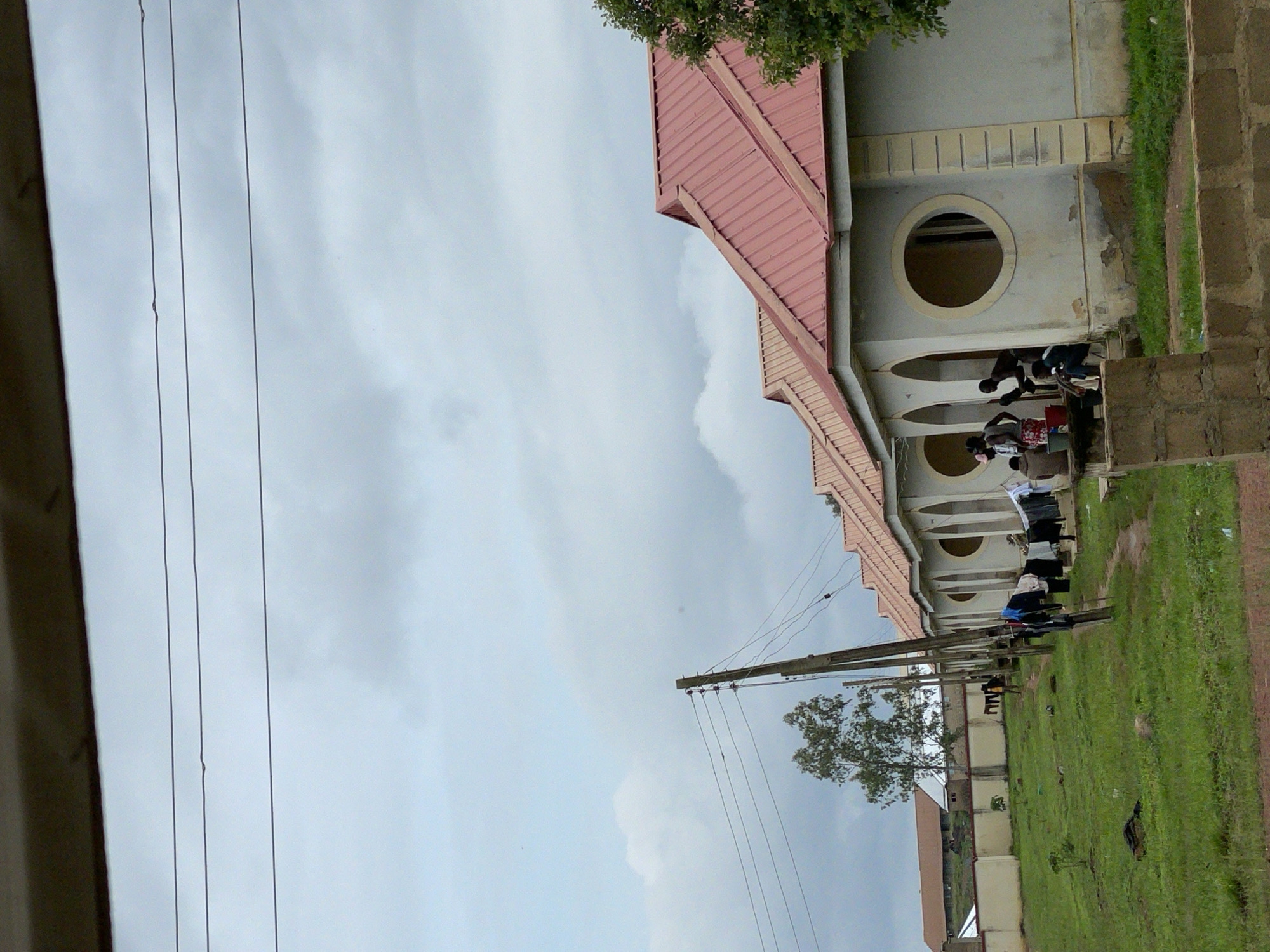 In Durumi, Nigeria, a row of identical, single-story buildings with light-colored walls and reddish-pink corrugated metal roofs is situated. Each building features arched doorways and large circular windows. Several people are visible, some standing by the doorways and others seated on concrete ledges. Laundry is hung on clotheslines strung between poles in front of the buildings. A grassy area with scattered debris is in the foreground, and a paved pathway leads towards the structures. In the background, a longer building with a similar roof is visible behind a low concrete wall. Electrical poles and wires traverse the scene, and a lone tree stands to the left. The sky is overcast with gray clouds.