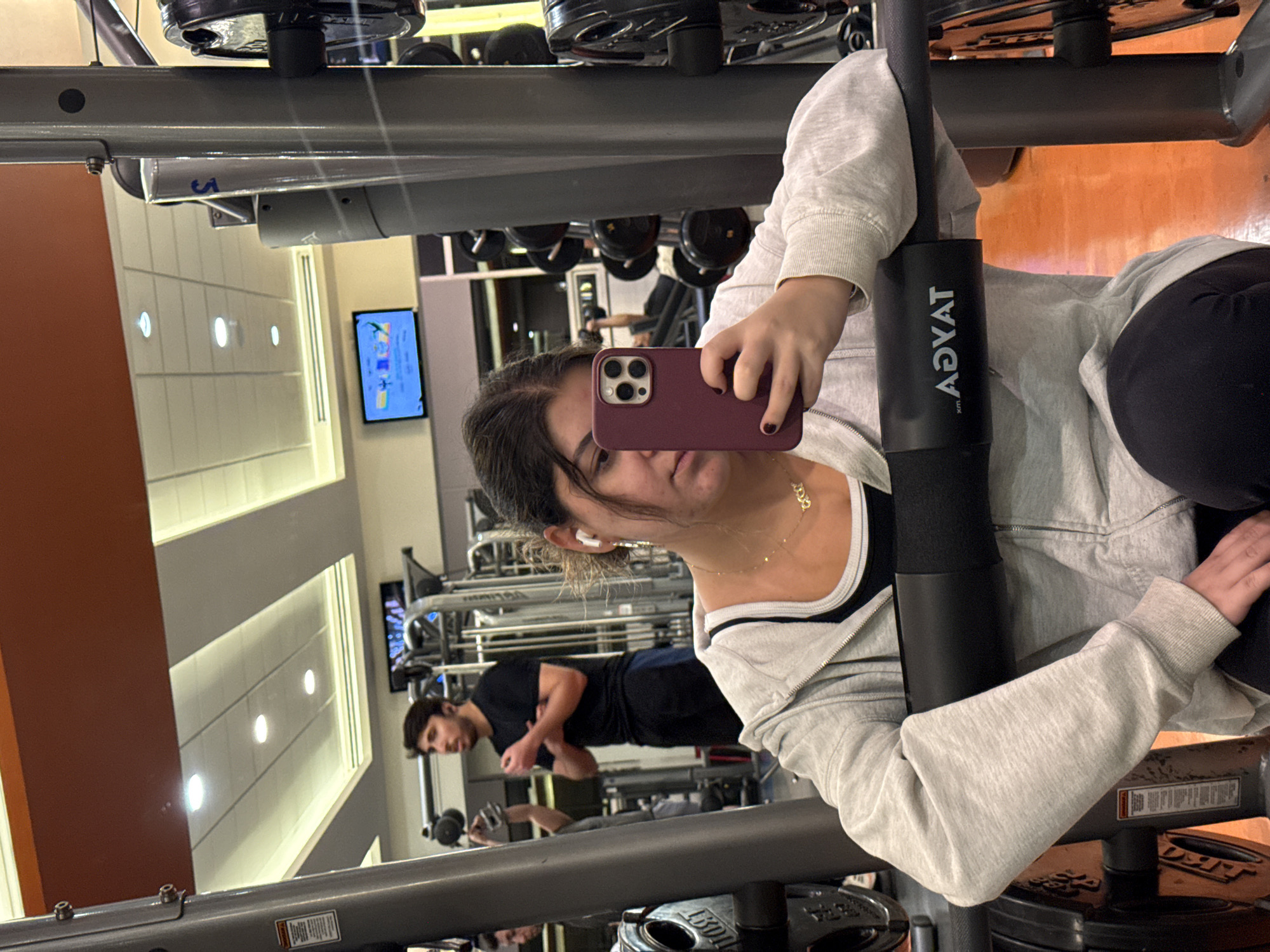 The image shows a woman taking a selfie in a gym. She is holding a maroon-colored smartphone up to her face. She is wearing a light gray zip-up hoodie over a black and white top. The gym contains weight machines and free weights. A man in a black shirt is visible in the background, lifting weights. A television screen is mounted on the wall in the background. The equipment in the foreground has the brand name "Advat" printed on it. The location is likely a gym in Mexico City, Mexico.