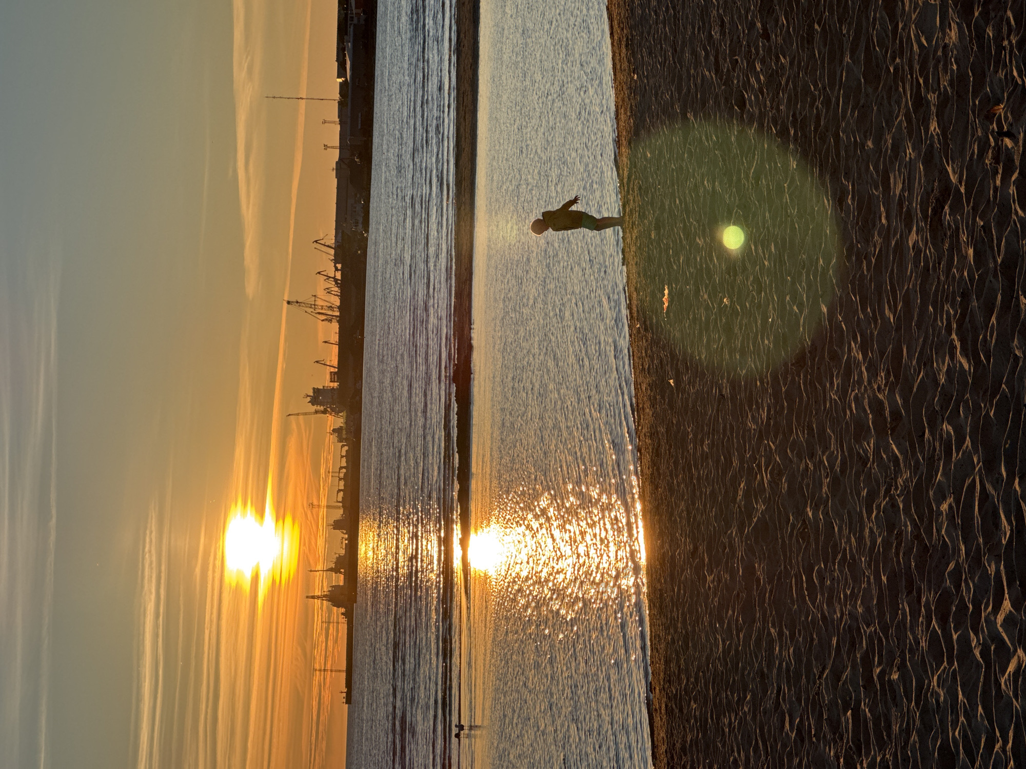 This is a vertical, eye-level, medium shot of a person standing on a sandy beach next to the water at sunset. The sun is low on the horizon, casting a warm, golden light and creating reflections on the water. The sky is a gradient of blue and orange, with some wispy clouds. In the distance, silhouetted against the sunset, is a port with cranes and buildings. The sand in the foreground has rippled patterns from the tide. There is a lens flare in the middle right of the image. The overall mood is peaceful and serene, evoking the end of a day at the beach. The location is likely Tallinn, Estonia, given the presence of a port.