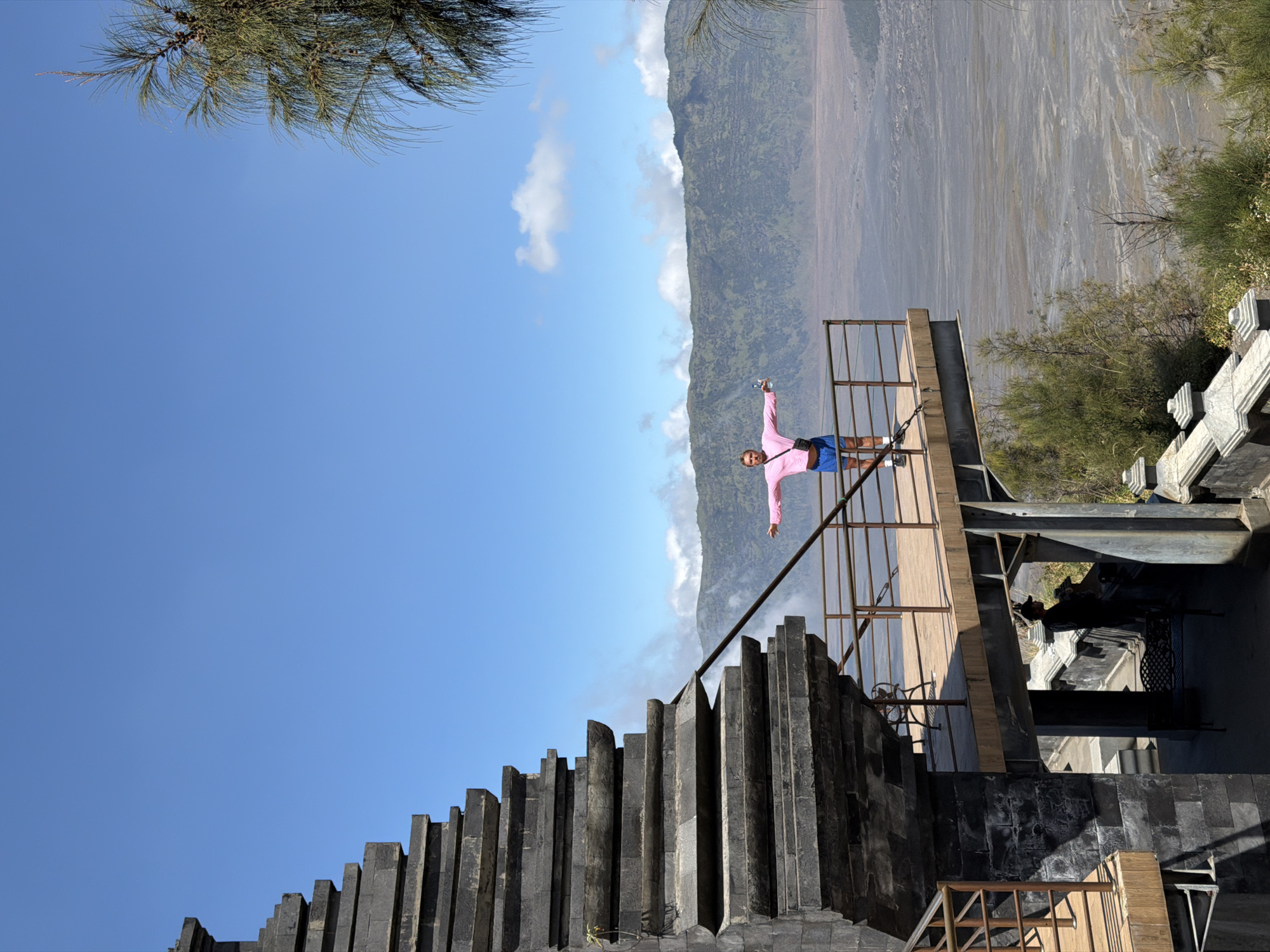 This image, taken in Cemorolawang, Indonesia, depicts a person standing on a viewing platform overlooking a vast, arid landscape. The platform, constructed with a wooden deck and metal railings, juts out from a stone structure with tiered steps, possibly part of a temple or monument. The person, wearing a pink long-sleeved shirt and blue shorts, stands with arms outstretched, holding a water bottle in their right hand. The background features rolling green mountains partially shrouded in mist, under a clear blue sky dotted with white clouds. Trees with feathery foliage frame the right side of the image. In the foreground, shadows suggest the presence of other structures and vegetation.