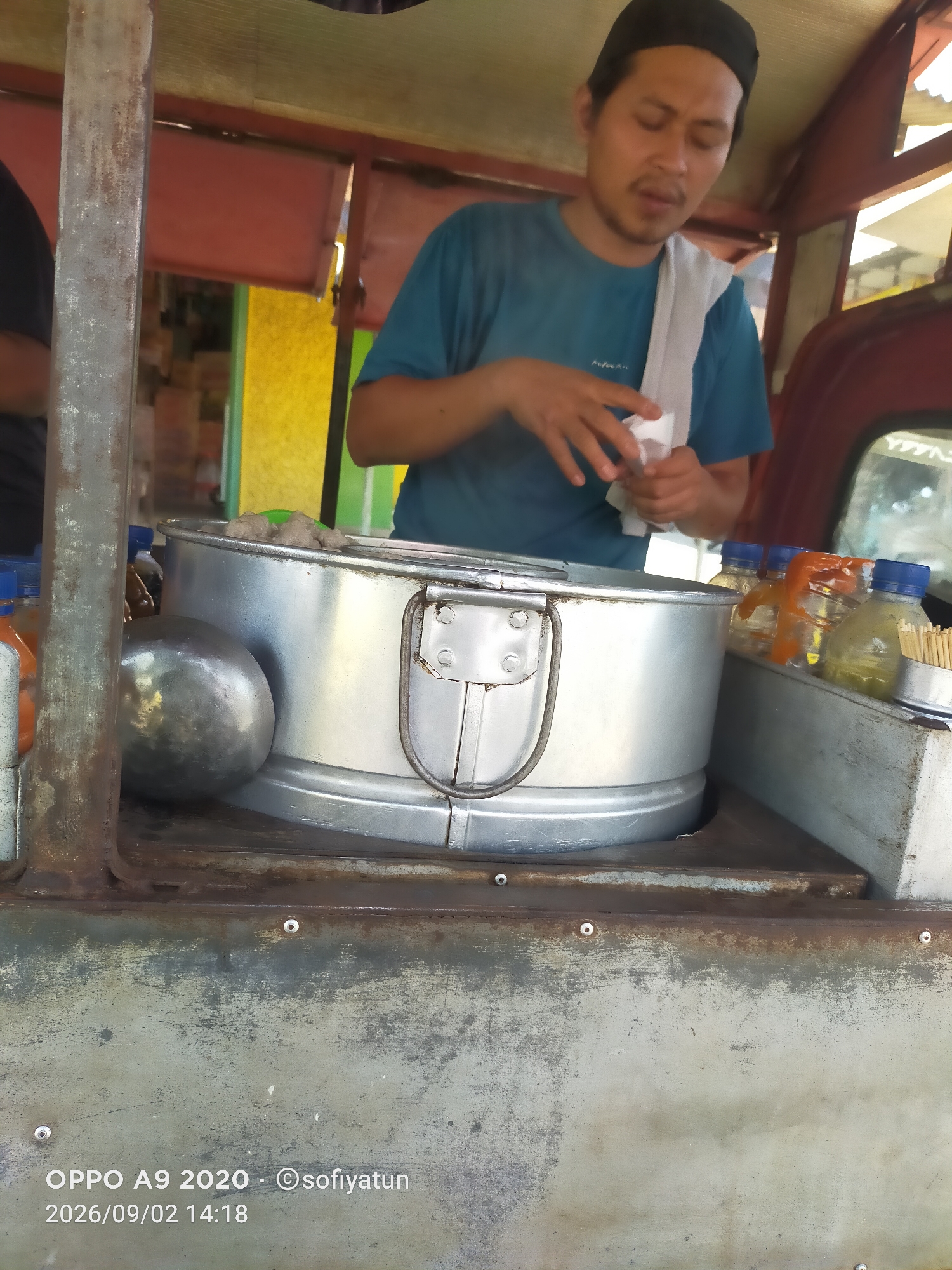 In the city of Tayu, Indonesia, a street vendor, a man wearing a blue t-shirt and a black cap, stands behind a food cart. He is holding a white paper napkin and appears to be preparing to serve or clean. The cart contains a large, metallic steaming pot filled with round, brown food items, possibly meatballs. Next to the pot, a metal ladle rests. To the right of the pot are several small plastic bottles filled with liquids, some with orange wrappers, and a container of toothpicks. The cart itself is made of metal and shows signs of wear and rust, particularly on the lower section where a date and time stamp "2026/09/02 14:18" and text "OPPO A9 2020 - sofiyatun" are overlaid. The background is blurred, but hints of colorful structures suggest an outdoor market or street setting.