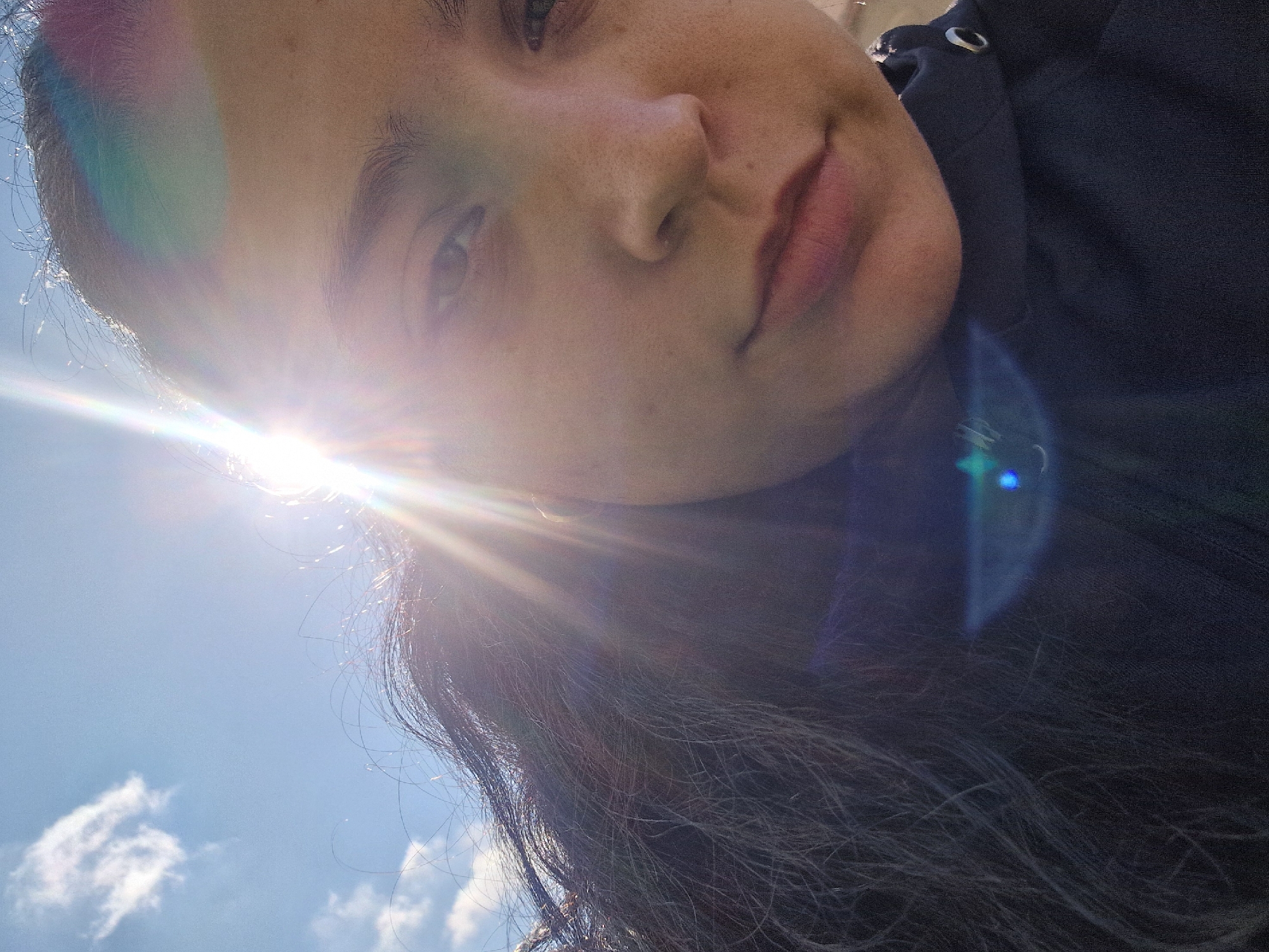 A close-up, tilted, medium shot depicts a person's face against a sky backdrop. The bright sun, positioned slightly below and to the left of the face, casts a significant glare, partially obscuring the face and creating lens flares. The person has dark hair that extends into the lower portion of the frame. The sky is a clear blue with some scattered white clouds visible in the lower left corner. A dark-colored garment with metallic detailing is visible on the right side of the image.
Location: Unknown, Unknown.