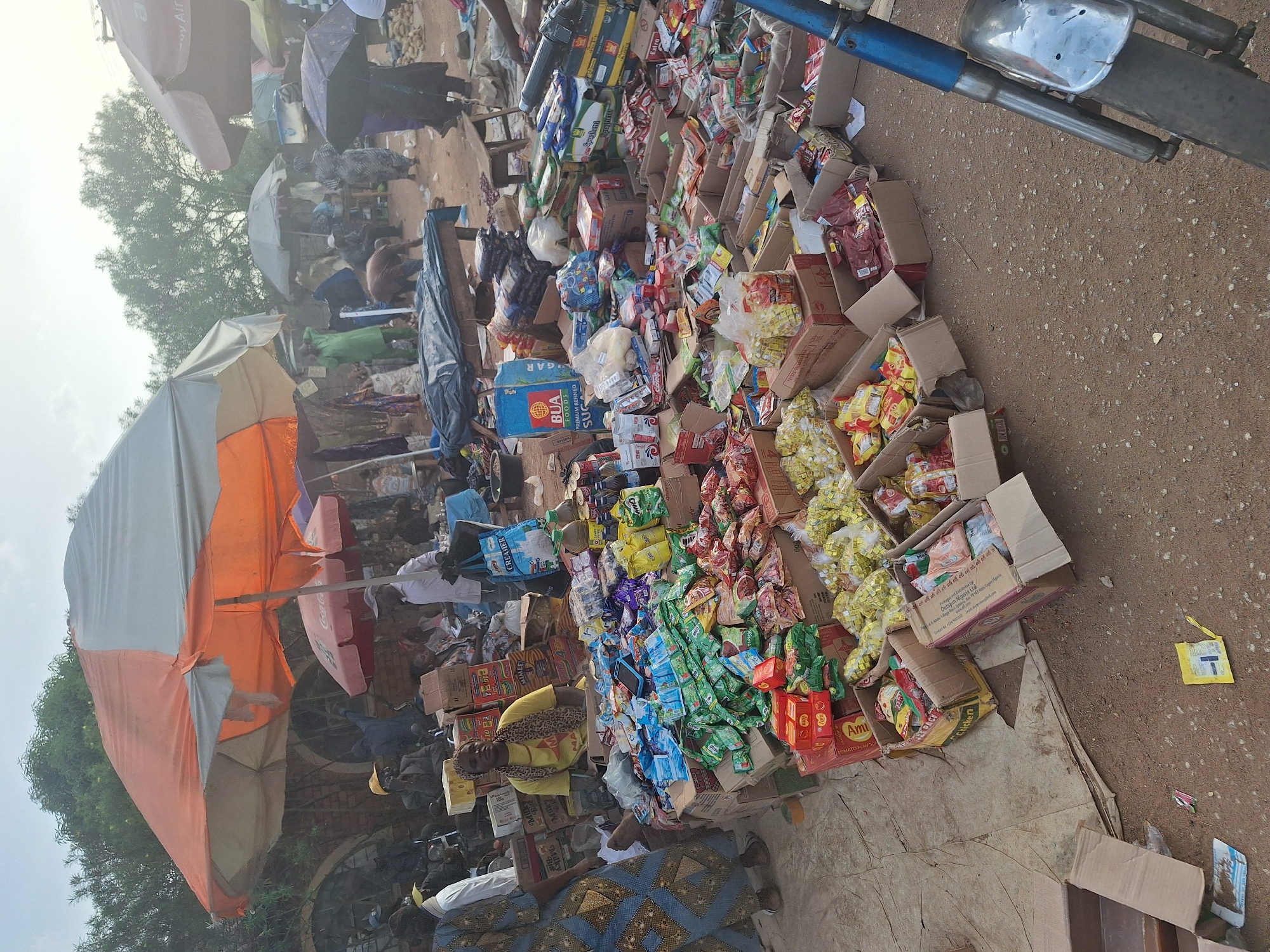 The image depicts a street market in Kakuri Gwari, Nigeria. A vendor sits amidst a large assortment of packaged goods, including food items such as spices, tomato paste, and sugar, arranged in cardboard boxes. The scene is filled with other vendors and customers, some using umbrellas for shade. The ground is a mix of dirt and pavement. The front of a motorcycle is visible in the upper right corner.