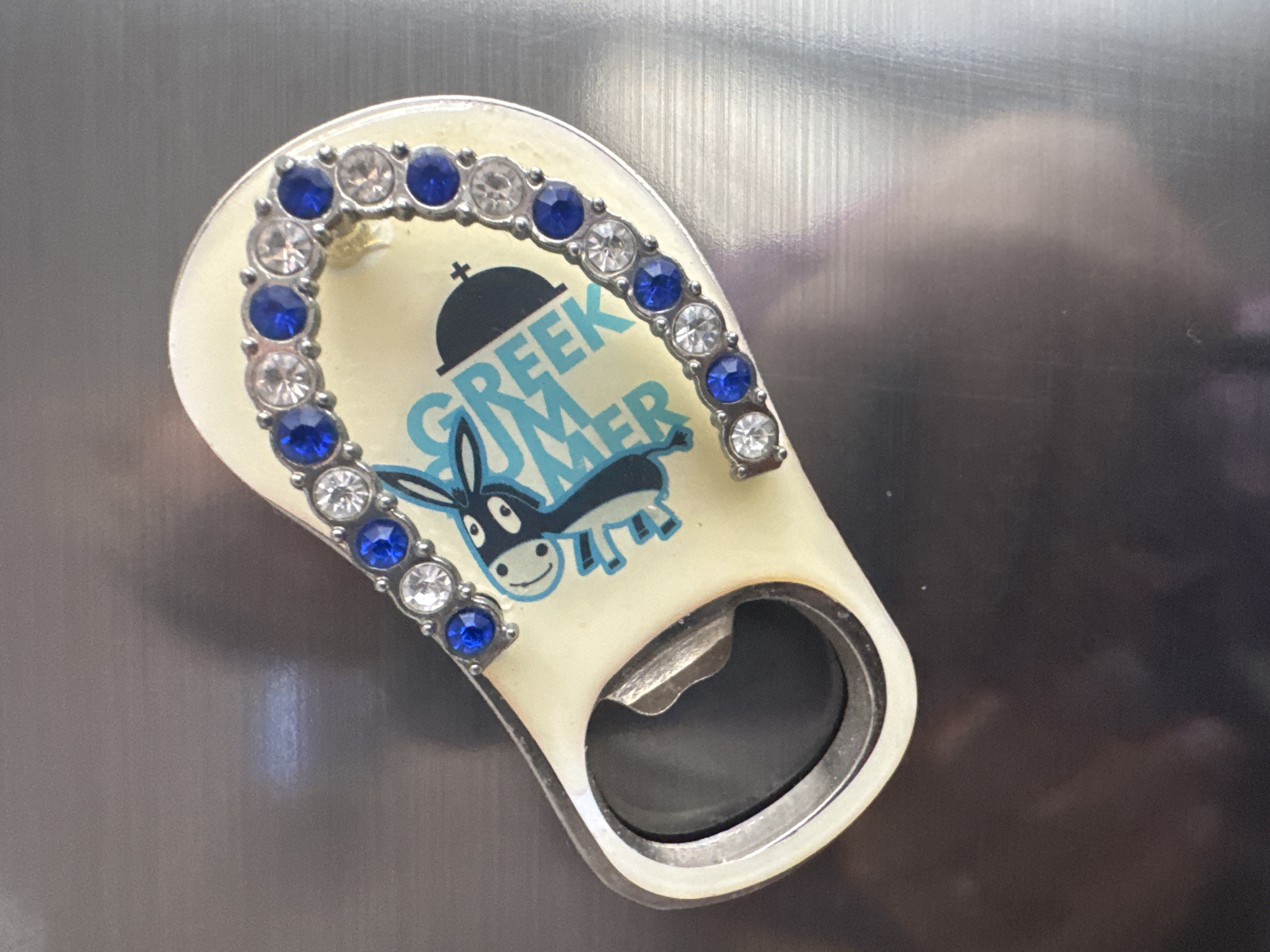 This is a close-up, overhead shot of a novelty bottle opener with a cream-colored handle decorated with a cartoon donkey and the words "GREEK SUMMER" in blue lettering. The handle is adorned with a curved row of rhinestones and blue faux gems. The metallic bottle opener part is on the left side of the frame. The object is placed on a reflective, metallic surface, possibly a refrigerator or a stainless steel appliance. The background is out of focus and shows a dark, diffused reflection. The image quality is moderate with some blur and glare.
There is no information in the image that can be used to determine its location in the city of Novosibirsk, Russia.