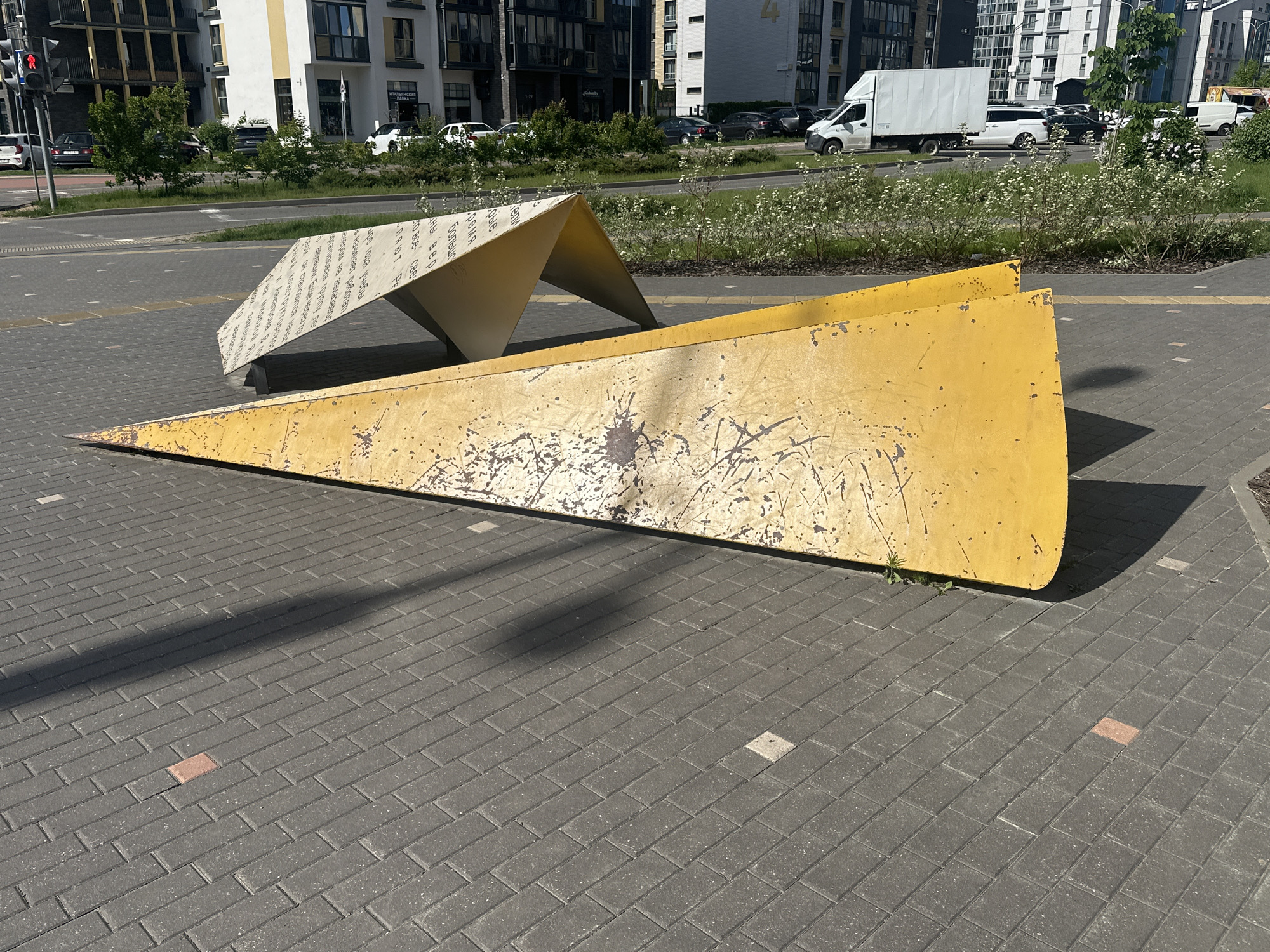 The image captures an outdoor scene in Бараўлянскі сельскі Савет, Belarus, focusing on two geometric sculptures positioned on a paved area.
In the foreground is a large, yellow, triangular structure. The surface is weathered, displaying scratches and imperfections. Behind it is a smaller, white sculpture with text on its surface.
The ground is paved with gray bricks, exhibiting a repetitive pattern. The paving is interspersed with a few randomly placed, lighter-colored square tiles and a red brick.
In the background, there is a street with a line of trees and green vegetation separating it from the sculptures. A row of modern apartment buildings is further back. Several vehicles are parked along the street, including a white delivery truck. The weather appears to be sunny, casting shadows from the sculptures onto the pavement. The sky is not visible in the image.
