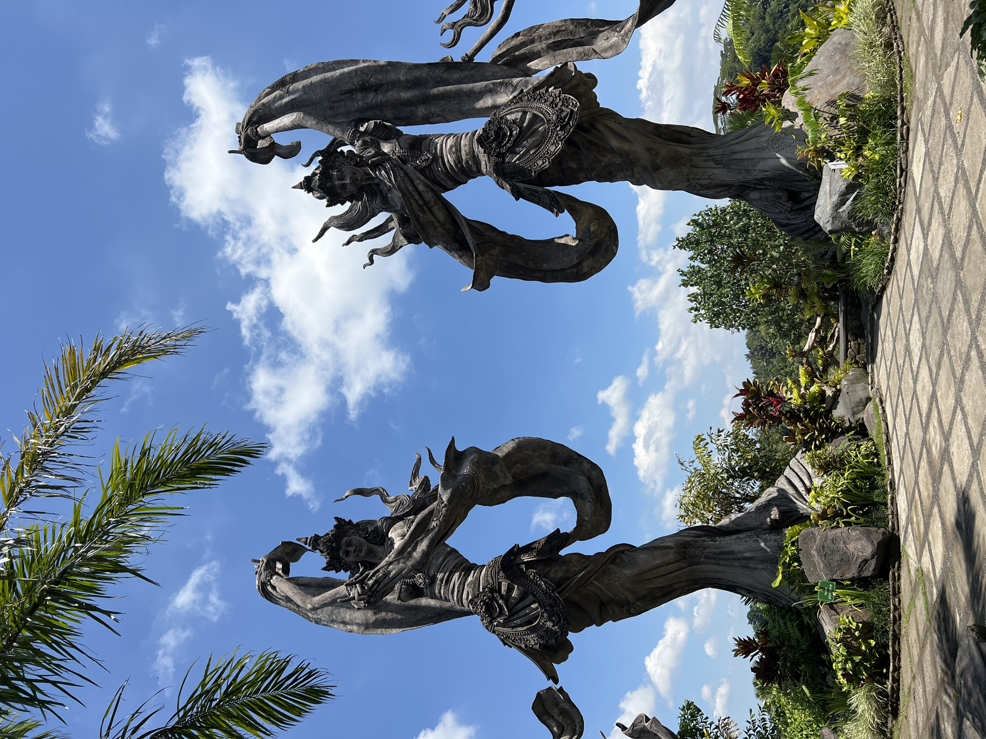 The image shows two gray statues of what appear to be Balinese dancers, set against a backdrop of a blue sky with scattered clouds. Palm trees are visible to the left of the image. The statues stand within a garden setting, with a gray tiled pathway and various greenery surrounding them. The location is in Kedewatan, Indonesia.