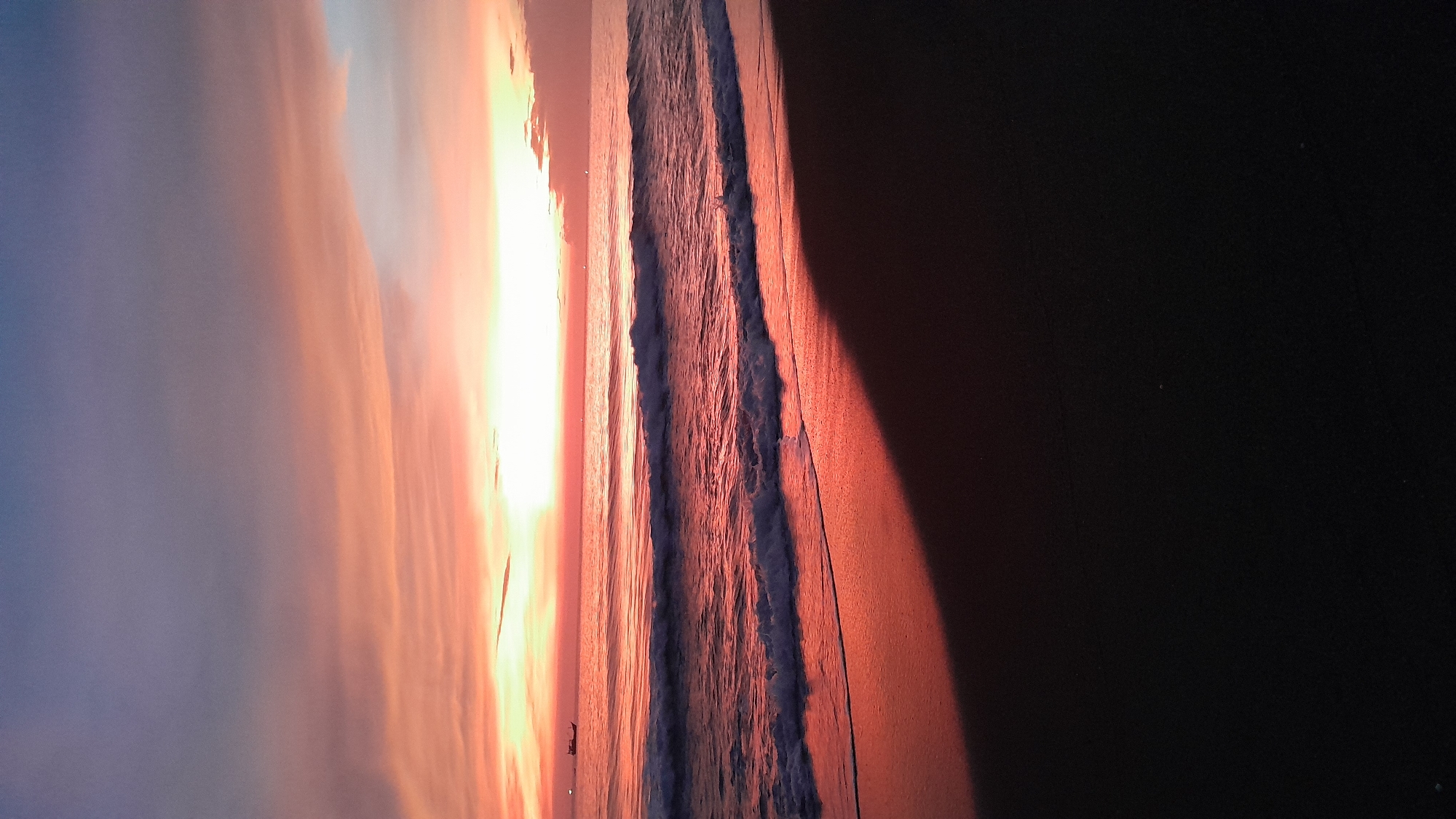 This image captures a coastal scene at either sunrise or sunset in Lhokseumawe, Indonesia. The sky displays a gradient from dark blue-grey at the top, transitioning through purple and pink, to vivid orange and red along the horizon, with several linear cloud formations. A bright light source, indicative of the sun, is prominent at the horizon line, casting a strong reddish-orange hue across the water. The ocean surface reflects these warm colors, showing multiple waves breaking towards the shore, with visible white foam at their crests. A dark silhouette of a traditional fishing boat is positioned on the left side of the horizon. The foreground consists of dark, wet sand on a beach, reflecting the reddish light where it meets the water. A distinct dark shadow or area is present in the lower right portion of the sand. No individuals are depicted in the scene.