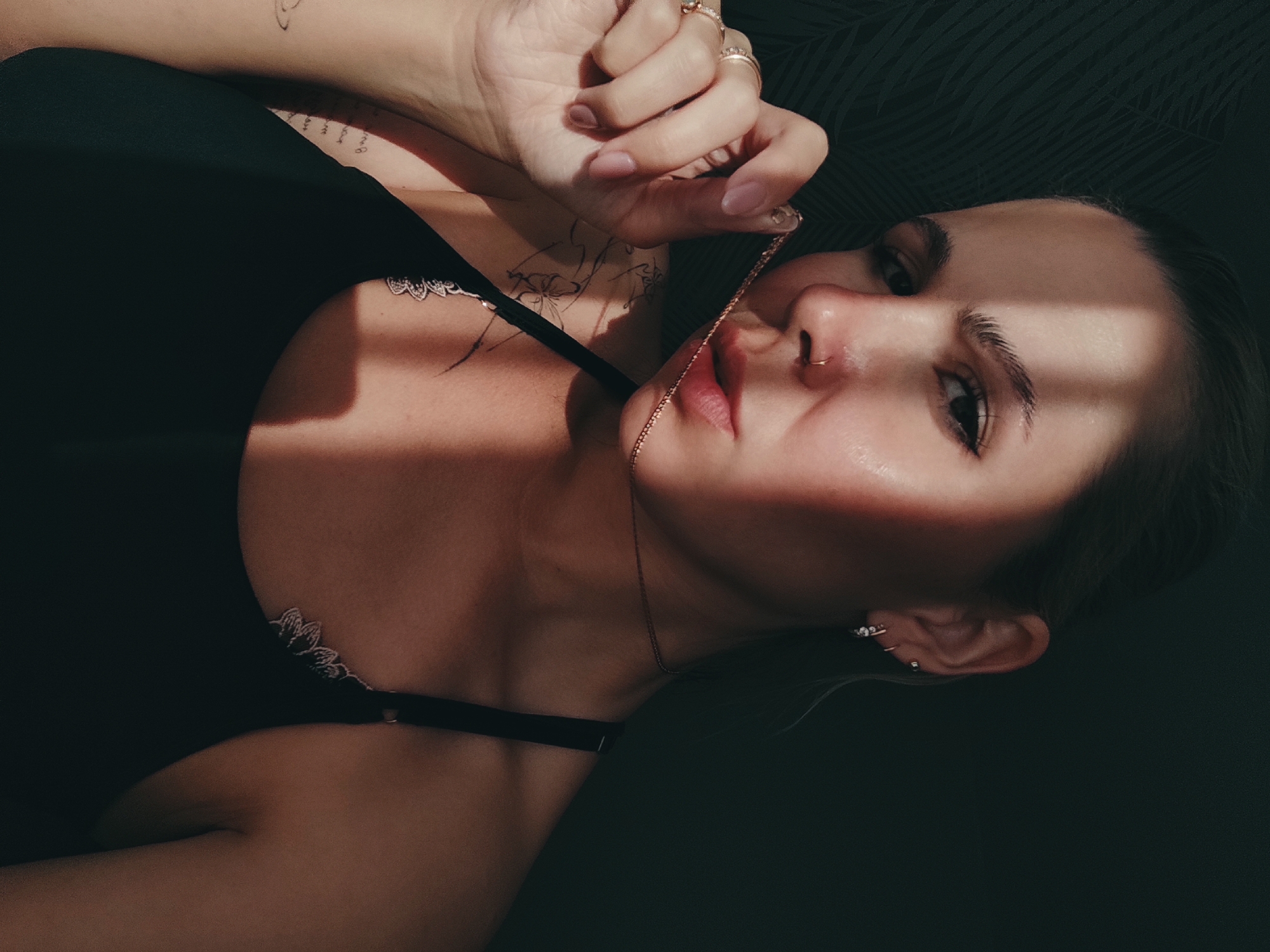 A woman is depicted from a low-angle perspective. She lies on what appears to be a dark surface or a black object. She is wearing a black top with a floral-patterned lace trim. Her right hand holds a thin gold chain necklace in front of her face. She has rings on her fingers and multiple ear piercings. A nose ring is visible. She has tattoos on her upper chest and arm. A beam of sunlight falls across her face, creating a strong contrast. The background is dark with a subtle, possibly leafy, pattern. Location information is not possible based on the image provided.