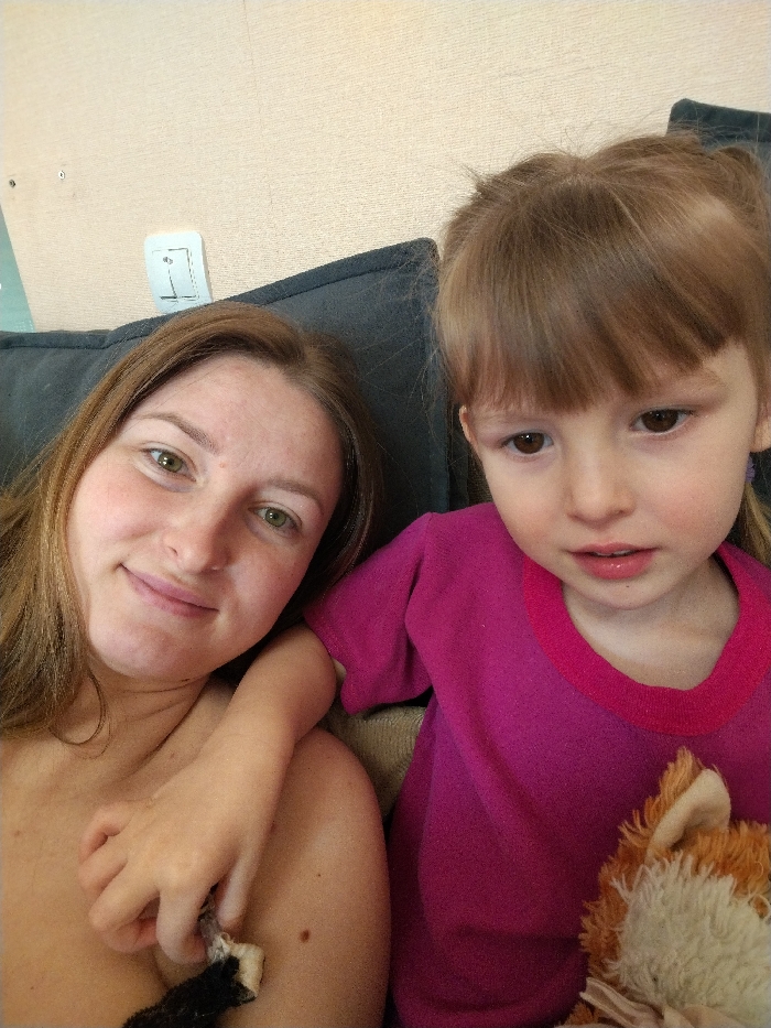A woman with light brown hair and fair skin smiles slightly. Beside her sits a young girl with bangs, wearing a magenta shirt and holding a stuffed fox. The woman's arm is draped around the child. A dark gray pillow or cushion is behind them. The background wall is a light beige color with a white electrical outlet.