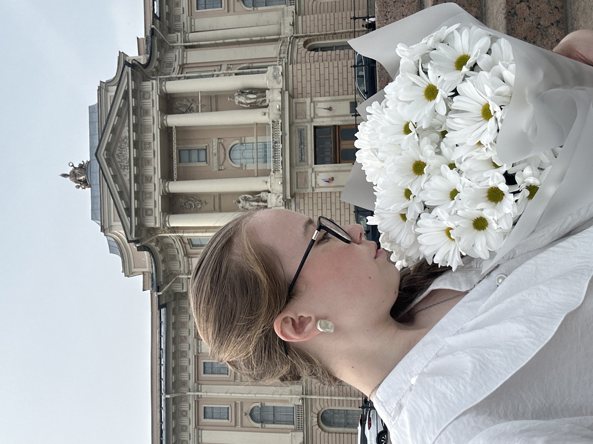 A medium shot depicts a woman holding a bouquet of white daisies in front of a building in Saint Petersburg, Russia. She wears glasses and a white shirt. The building behind her has columns and statues. The sky is overcast.