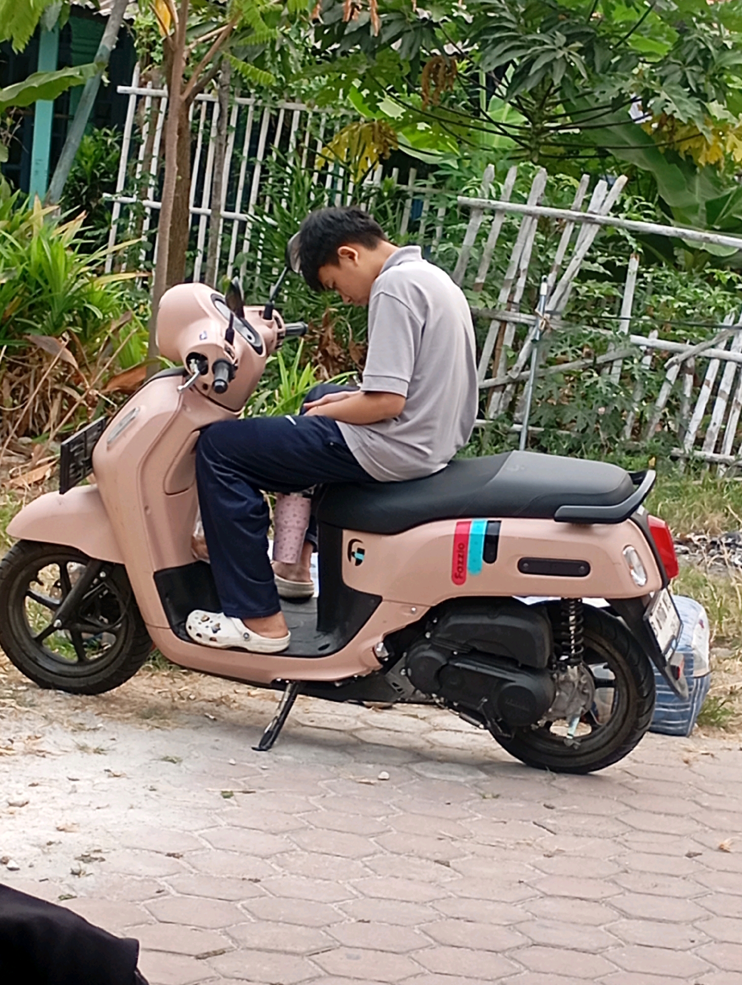 A young man wearing a grey t-shirt and dark blue pants is sitting on a light pink scooter. He is looking down at his phone. The scooter has a black seat and black wheels. There is a white plastic bag with blue stripes behind the scooter. The scene is outdoors, with lush green foliage and a white picket fence in the background. The ground is paved with hexagonal bricks. The scooter appears to be parked on a dirt path adjacent to the brick paving. The lighting suggests it is daytime. The location is identified as Babelan, Indonesia.