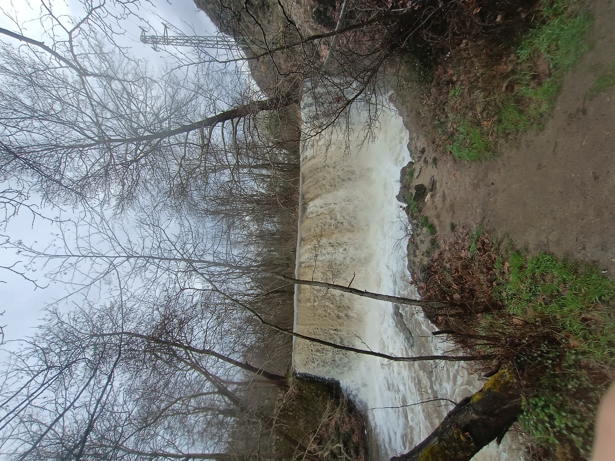 A landscape-oriented photograph shows a waterfall scene in Parvenets, Bulgaria. The image appears to be taken on an overcast day, with grey skies visible above bare tree branches. A structure with a lattice framework is partially visible through the trees in the upper left corner. The waterfall cascades over a straight edge and into a churning body of water below. The water has a muddy, brownish tint. The foreground includes bare trees and a patch of green vegetation on the right side. A dirt path runs alongside the waterfall on the right. The image is rotated 90 degrees counter-clockwise.
