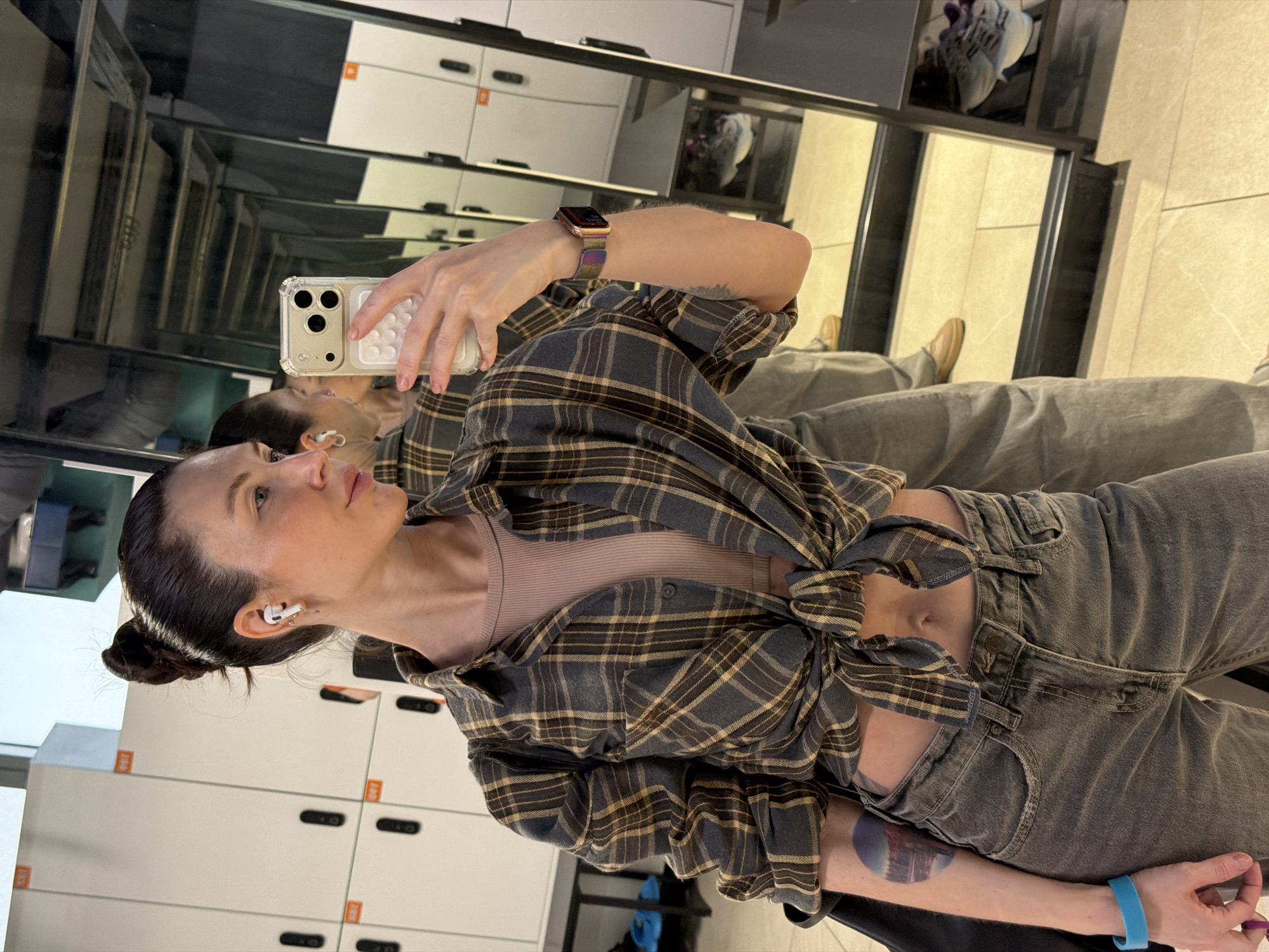 A woman is taking a selfie in front of a mirrored wall. She is wearing grey jeans, a mauve-colored tank top, and a dark grey flannel shirt tied at the waist. The woman has her hair in a bun and is wearing earbuds. She is holding a smartphone with a clear case. Her right arm has tattoos and a blue wristband. She also wears an Apple watch on her left wrist. Behind her are rows of white lockers with black handles and orange square details. The location is likely a gym or fitness facility in Perm, Russia, given the locker room setting. Her reflection is visible in the mirrored wall, showing the full outfit and shoes.