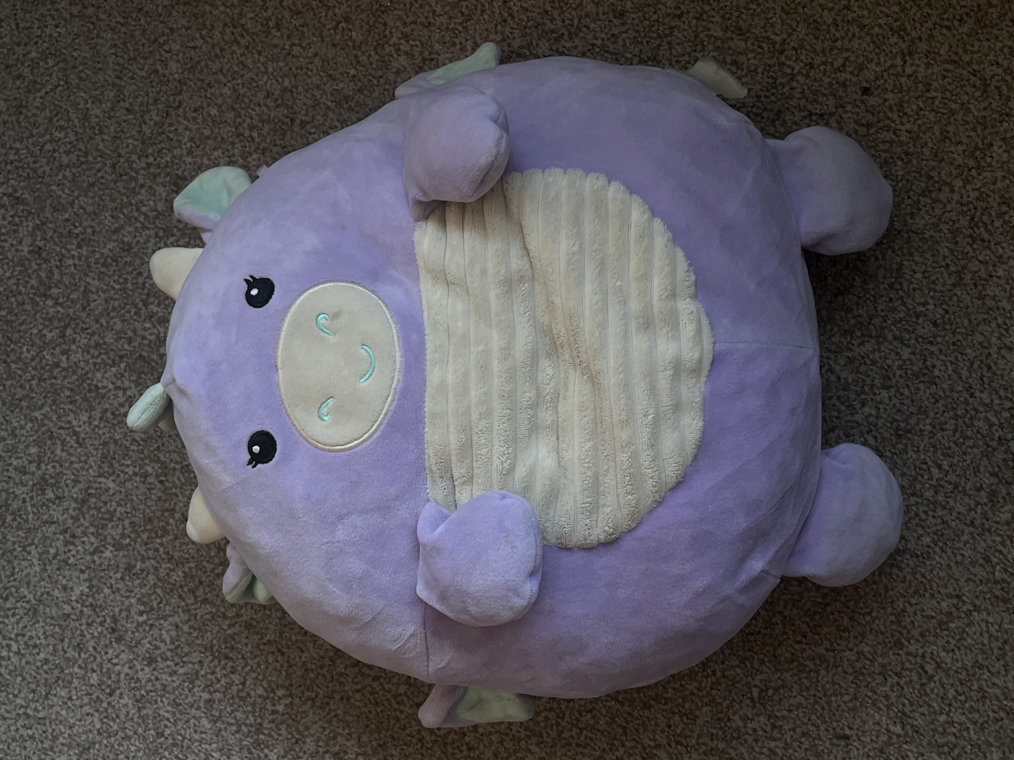 A lavender plush dragon toy lies on a brown carpet. The dragon has a round body, short arms and legs, and small wings on its sides and head. Its face features black eyes, a light tan snout with two small light blue curved lines for a smile, and two small light blue crescent shapes above its eyes. The dragon's belly is a textured light tan color. The image is taken from a top-down perspective. This scene is located in Genemuiden, Netherlands.