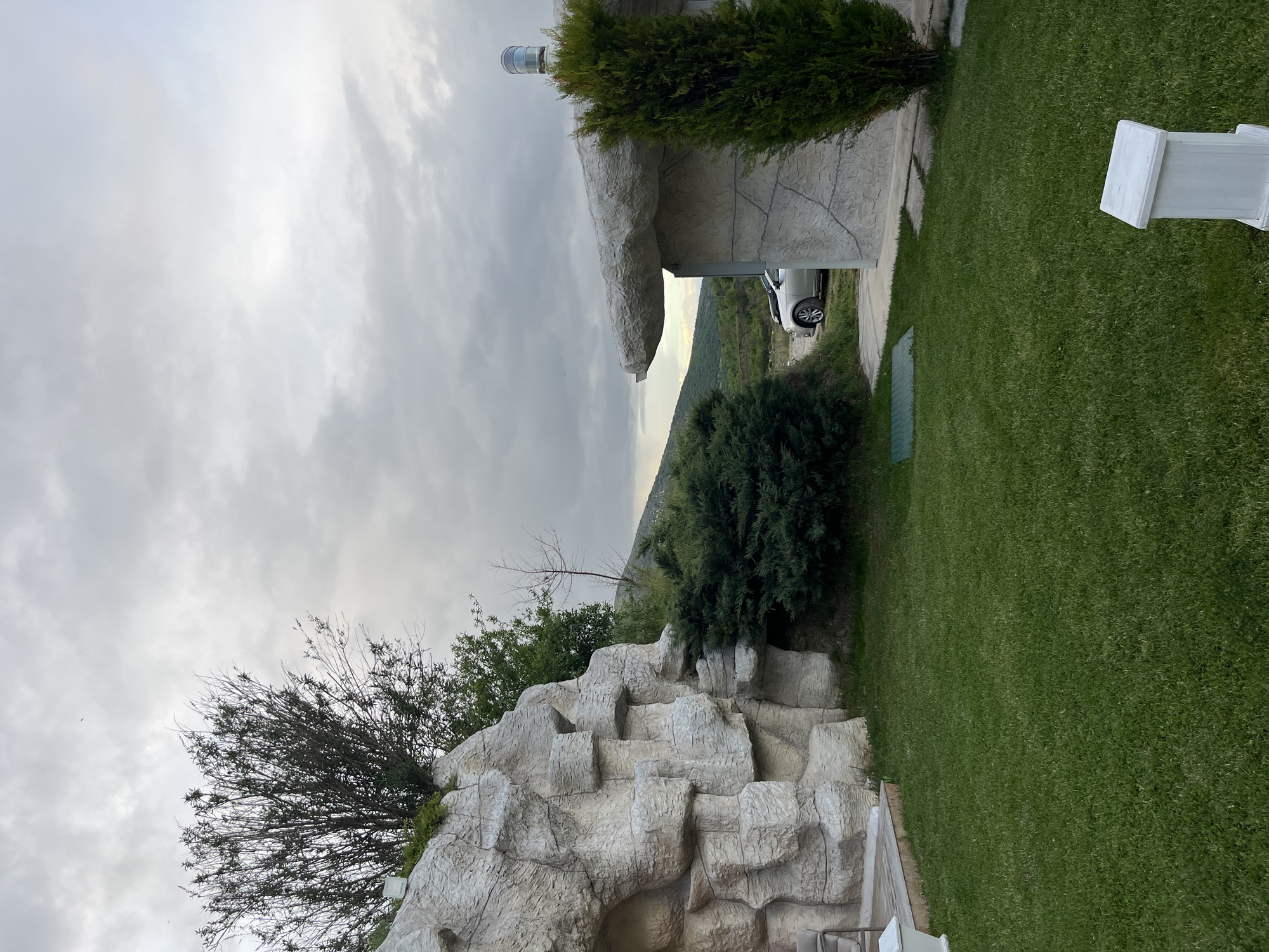 The image depicts a landscape with architectural features, possibly in Koprivlen, Bulgaria. In the foreground, there is a structure resembling a rocky outcrop with vegetation growing on it. Adjacent to this is a green lawn, and a portion of a white fence. A building with a simulated rock exterior and a tree growing on its roofline is situated further into the frame. A white vehicle is parked under the shelter of the building. The background features a hillside, and the sky is overcast with grey clouds. The orientation of the image is rotated.