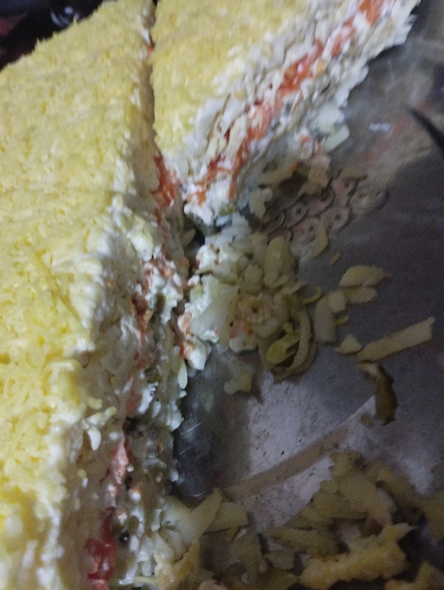 A layered salad, possibly a Mimosa salad given its appearance of shredded egg and other ingredients, is shown close-up. The salad is partially cut, revealing its distinct layers of yellow (likely shredded egg yolk), white (possibly shredded egg white or mayonnaise), and a reddish layer (potentially carrots or other vegetables). The salad is placed on a metallic surface with some scattered, peeled vegetables or salad ingredients around it. The lighting is dim and the image is slightly out of focus, making it difficult to discern fine details of the food. There is no indication of the city of Bilajari, Azerbaijan, or any people present in the image.