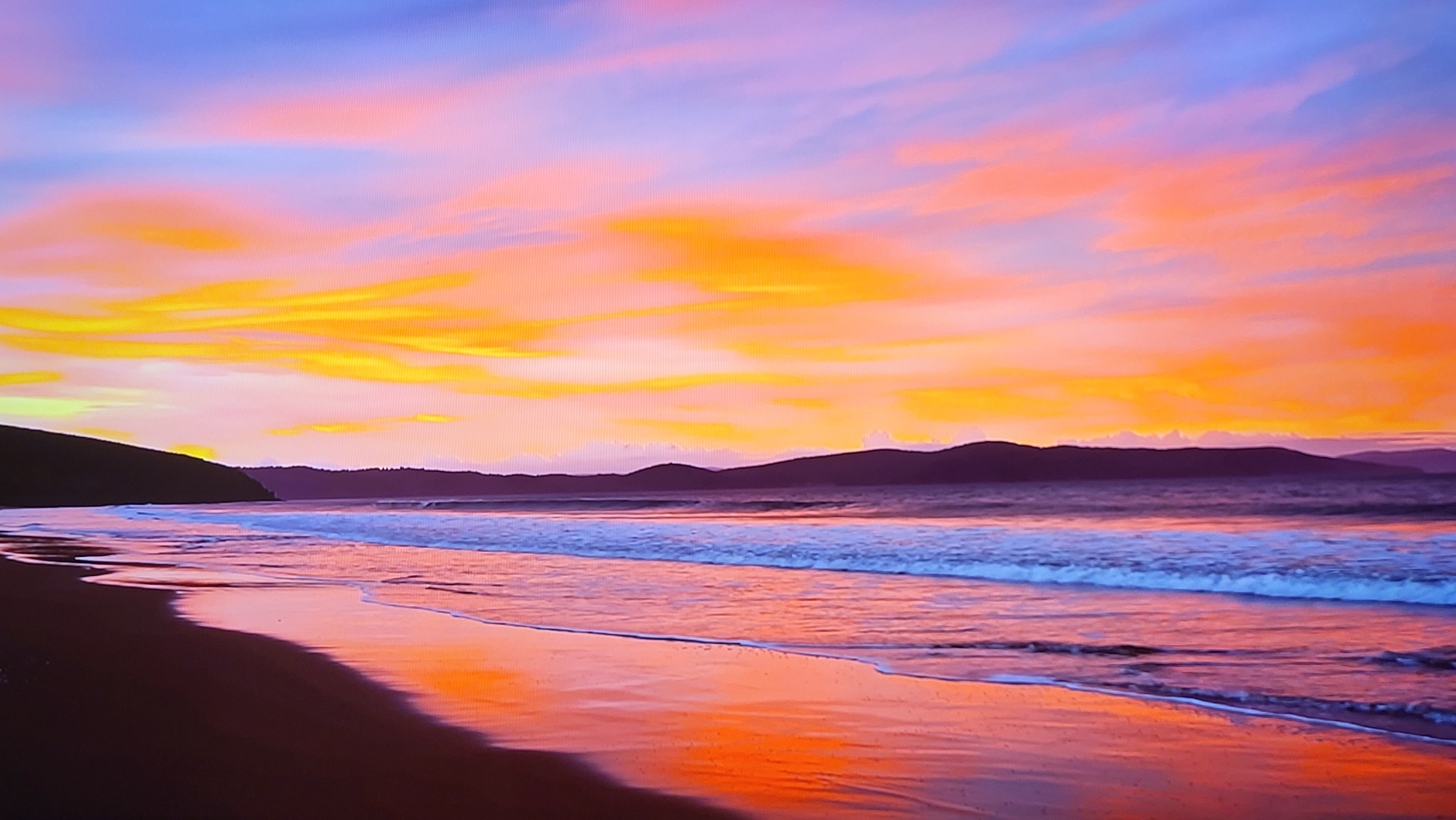 The image is a vibrant and colorful photograph of a beach at sunset. The sky is ablaze with orange, yellow, pink, and purple hues, which are also reflected in the wet sand of the beach. In the distance, there are dark silhouettes of hills or mountains. Gentle waves are rolling onto the shore. The beach is dark in the foreground, appearing almost black in some areas. The image contains no people or notable objects beyond natural elements. There is no information to indicate this photograph was taken in Yerevan, Armenia.