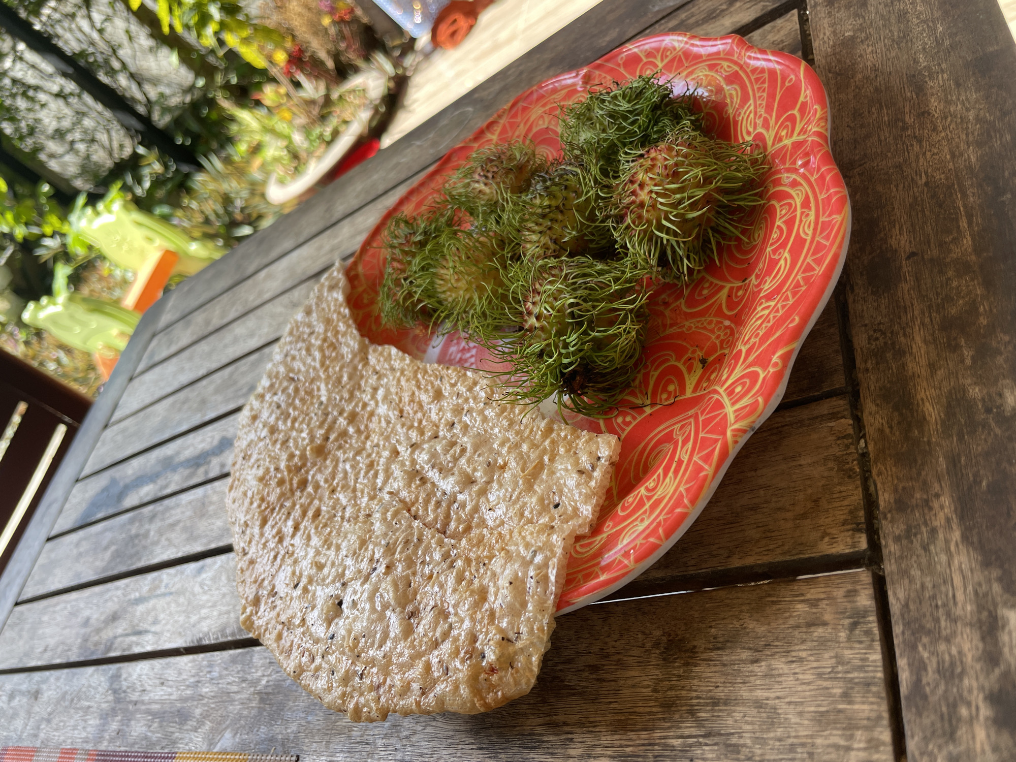 A single, round, textured cracker rests on a red and gold patterned plate, partially overlapping a cluster of green, spiky rambutans on a wooden table. The scene is outdoors, with a blurred background of green foliage and what appears to be outdoor furniture. The lighting suggests a sunny day. The image is shot from a slightly elevated angle, looking down on the table. No people are visible. The location is identified as Phu Thuy Ward, Vietnam.
