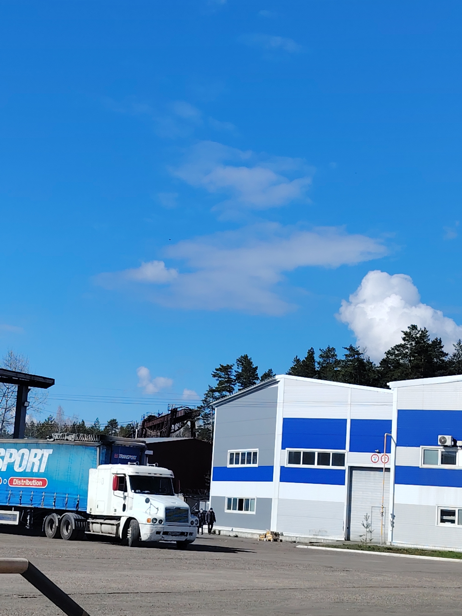 A truck with a blue trailer is parked on a paved surface in front of two industrial buildings with white and blue facades. The trailer has the word "TRANSPORT" printed on its side. A few people are walking nearby. A gantry crane is visible behind the truck and buildings. In the background, there is a line of trees and a blue sky with scattered clouds. The location is Noviy, Russia.