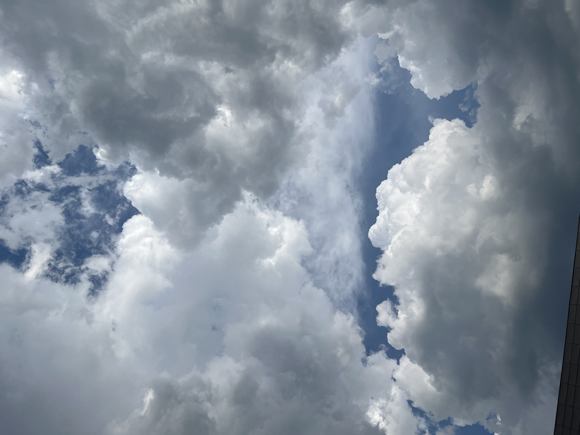 A wide-angle, low-angle shot captures a sky filled with dramatic, voluminous cumulus clouds, varying in shade from bright white to deep gray. Patches of clear blue sky are visible between the clouds, particularly towards the top of the frame. A dark, angular structure, possibly a building's rooftop or facade, runs along the bottom edge of the image, suggesting an urban setting. The lighting indicates it is daytime, possibly late afternoon, given the contrast between the bright and shadowed parts of the clouds. The image does not contain any people or specific actions. The scene is purely a skyward view, devoid of any identifiable landmarks that would place it in a specific city.