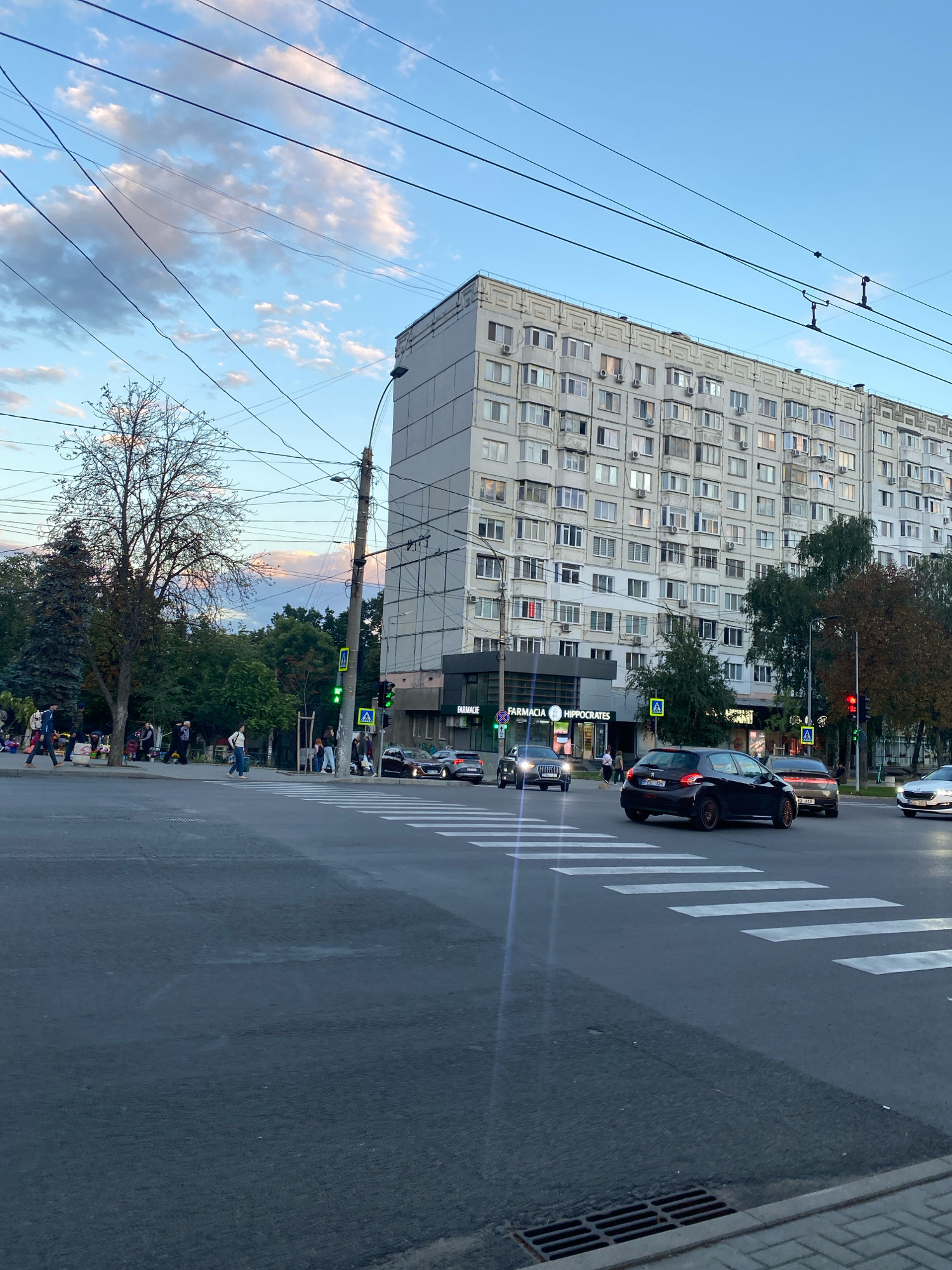 The image depicts a street intersection in Chișinău, Moldova. A large, multi-story apartment building dominates the background, with numerous windows and air conditioning units visible. Overhead power lines crisscross the sky. Several cars are present at the intersection, including a dark-colored hatchback in the foreground, an Audi SUV, and a silver car to the right. Pedestrians are seen on the sidewalks and crossing the street at a marked crosswalk. One pedestrian in the foreground is wearing a white backpack. Traffic lights are visible, with some showing green and others red. Storefronts with signage, including "FARMACIA" and "HIPPOCRATES," are visible on the ground floor of the building. Trees line the street, some with autumn foliage. The sky shows a mix of blue and clouds, with hints of sunset colors. The overall scene suggests a typical urban setting during daytime, possibly late afternoon or early evening, given the lighting and sky conditions.