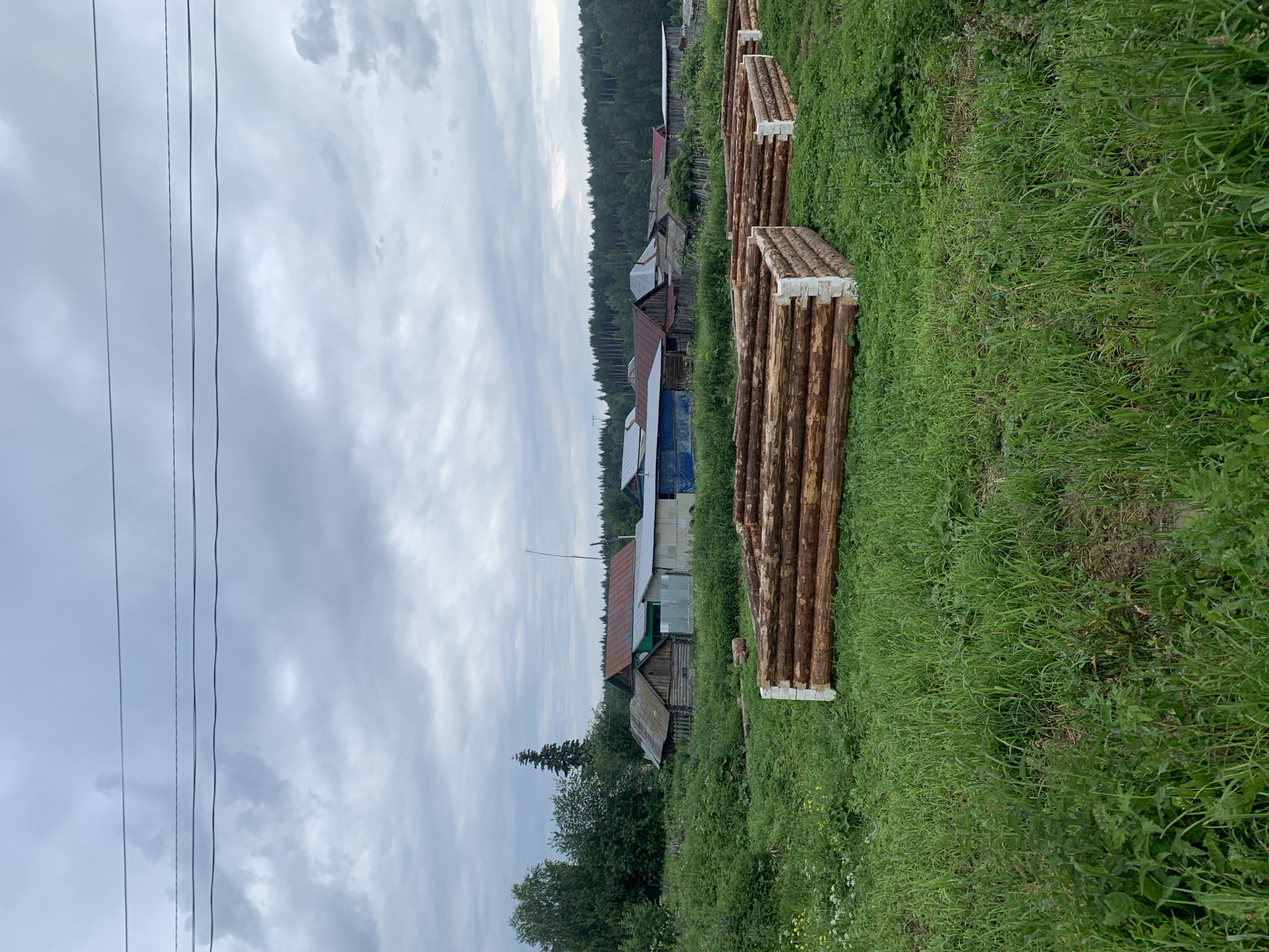 In a rural village setting in Barsai, Russia, a cloudy sky looms over a collection of rustic wooden structures. In the foreground, two unfinished log foundations, each several layers high and resting on white concrete blocks, sit amidst tall green grass. Beyond these foundations, a small village of houses with corrugated metal and wooden roofs is visible. Dense green trees and a forest line the horizon in the background, completing the pastoral landscape. Power lines stretch across the upper portion of the frame.
