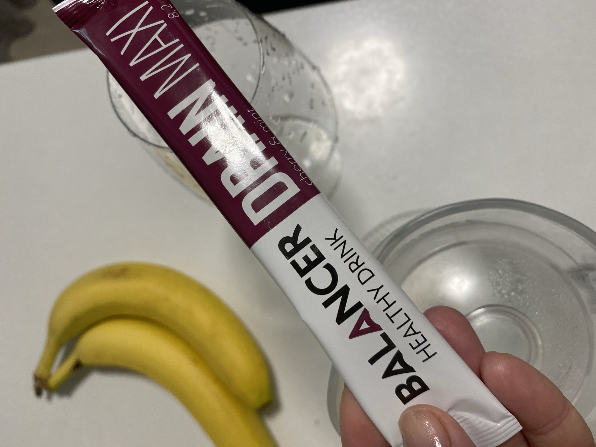 A hand holds a white and maroon single-serving packet labeled "BALANCER HEALTHY DRINK DRAIN MAXI" with "cherry & mint" also visible on the maroon section. Behind the packet is a mostly empty clear glass. In the lower left corner, there are two ripe bananas. The background is a plain white surface. Another clear glass or container sits partially visible to the right of the packet. The location is not apparent from the image.