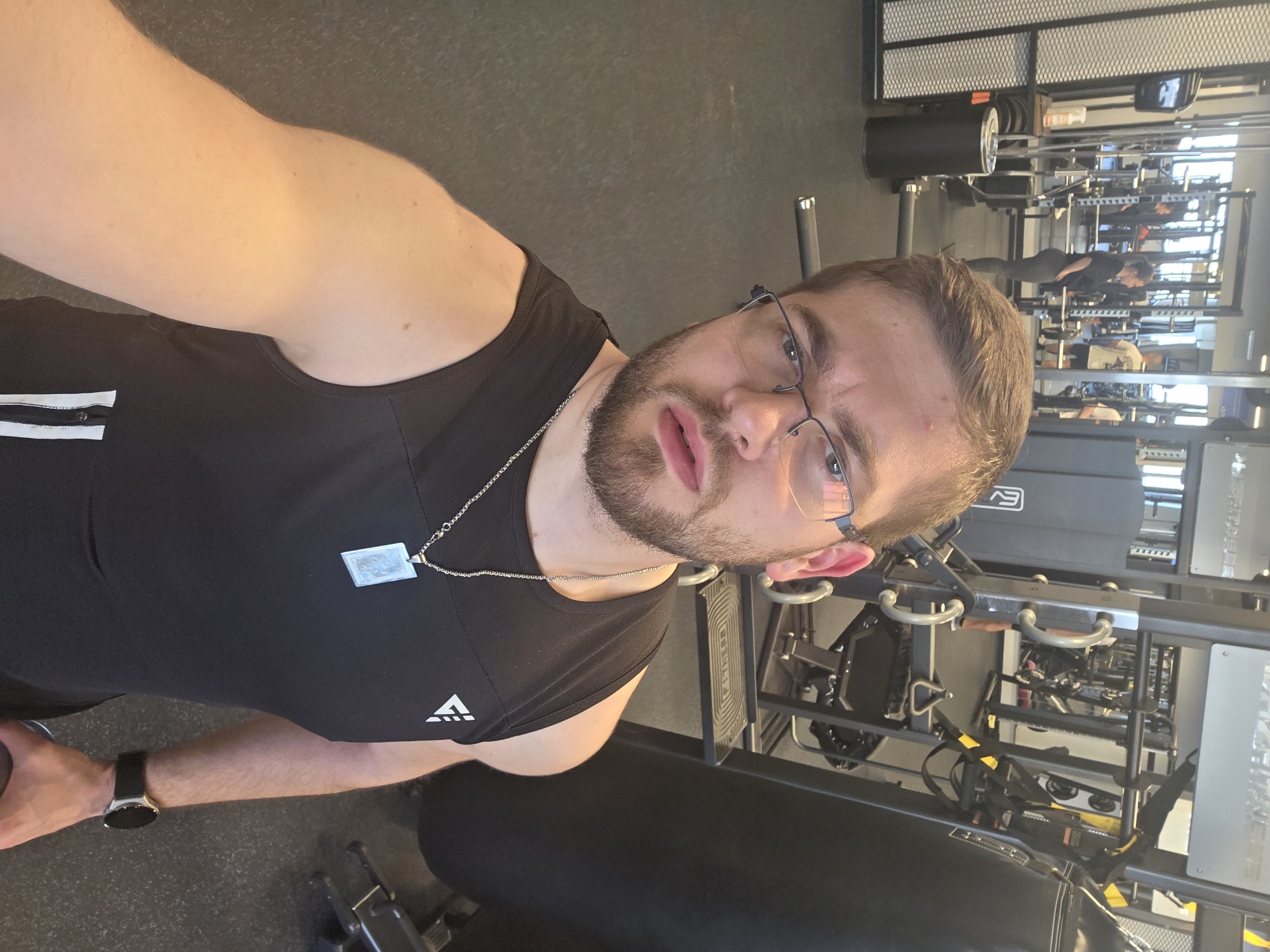 The image shows a man in a gym setting. He is wearing a black tank top, glasses, and a smartwatch. He has a chain with a pendant around his neck. The gym equipment is visible in the background. The location cannot be identified as Plovdiv, Bulgaria from the given information.