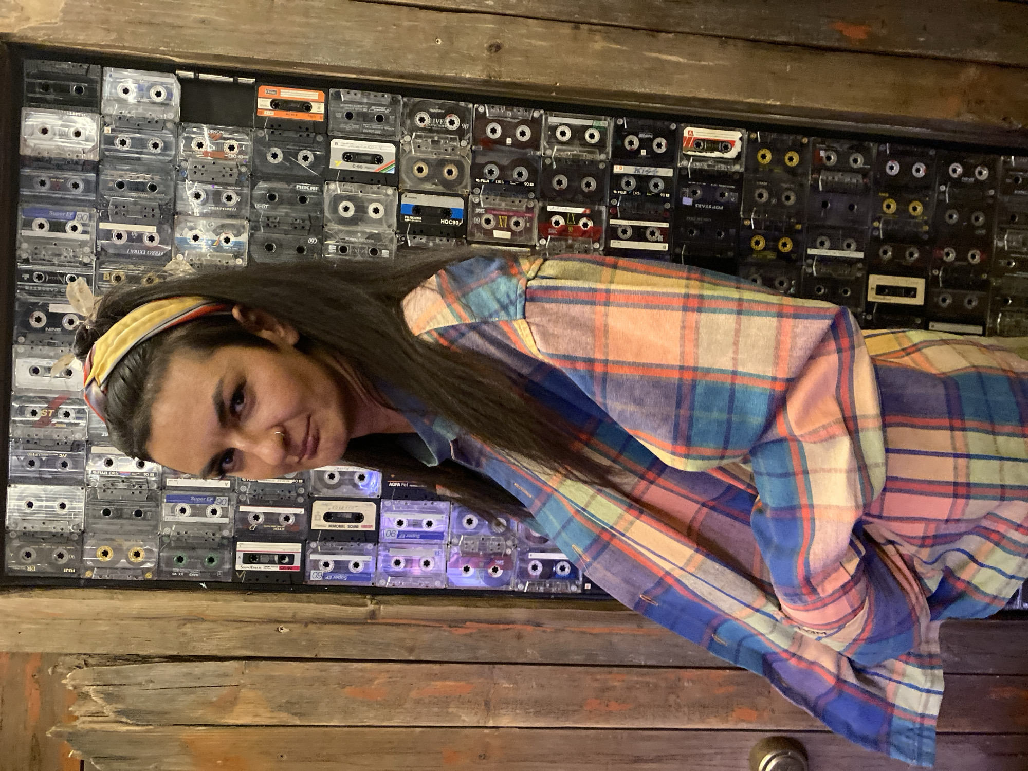 A woman with long dark hair, wearing a yellow headband and a colorful plaid shirt, is standing in front of a wall decorated with numerous audio cassette tapes. The tapes are arranged in rows within a wooden frame. The woman is looking at the camera with a slight smile. The environment appears to be an indoor setting, with wooden surfaces visible above and below the cassette tape display. The lighting is somewhat dim. There is no indication of the location being in Moscow, Russia.