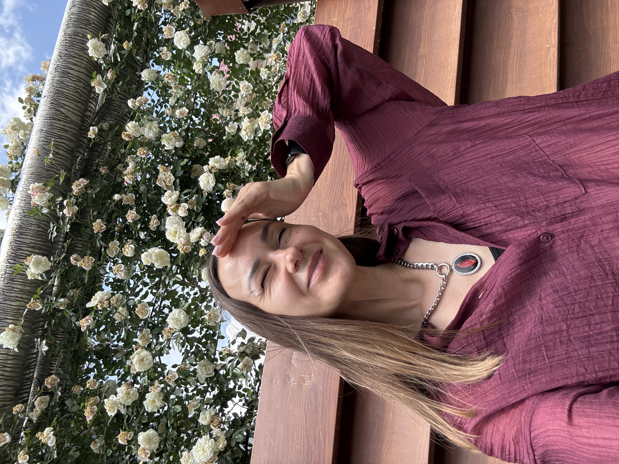 A woman is captured from the chest up, sitting on a wooden bench, with her right hand shielding her eyes from the sun. She is wearing a maroon, collared, long-sleeved shirt with a textured crinkled fabric and a chunky silver necklace with a circular pendant featuring a red stone. Her brown hair is long and falls over her left shoulder. Behind her is a dense trellis covered in white roses and green foliage. The sky is visible through the roses. The woman is squinting and smiling, suggesting a bright, sunny day. The wooden bench appears to be part of an outdoor seating area, possibly in a park or garden. The location is identified as Krasnaya Polyana, Russia.