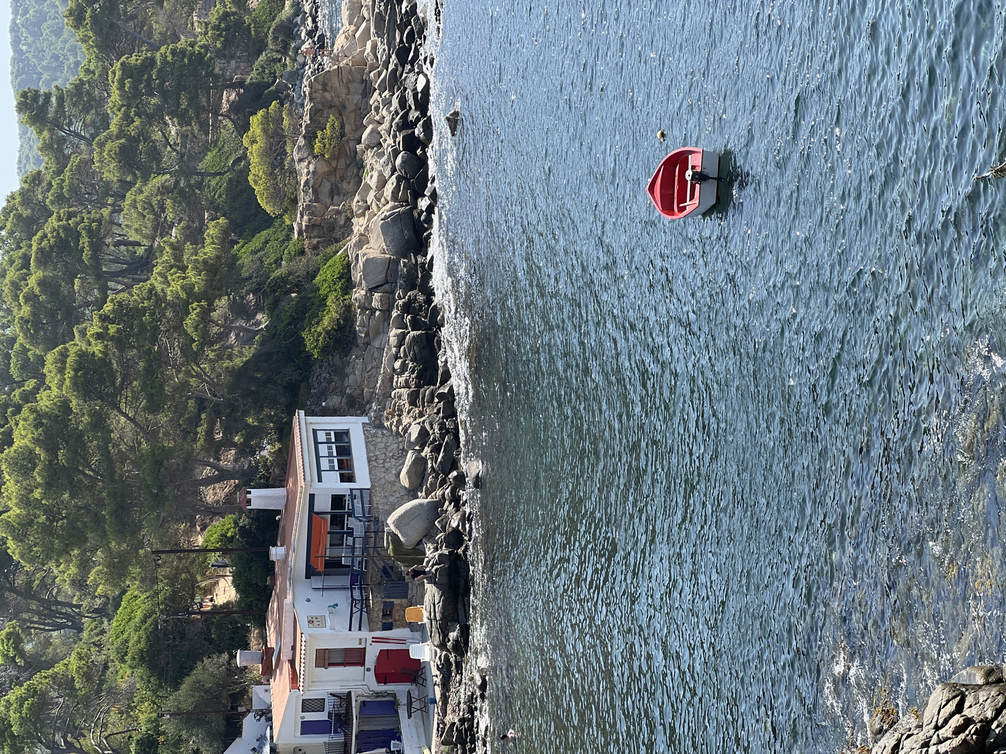 This is an eye-level, medium shot of a coastal scene in Palamós, Spain, on a sunny day. In the foreground, the deep blue water of the Mediterranean Sea is rippling, with sunlight glinting off its surface. A small, red and white motorboat is anchored in the middle of the water. In the midground, a rocky shoreline is visible, with large boulders and smaller rocks. A white building with a red-tiled roof is situated on the shore. The building has several windows, an orange awning, and a red door. There are tables and chairs outside the building. A person is seated on a rock near the building. In the background, a steep, tree-covered hill rises behind the building, with more pine trees covering the upper slopes. The sky is a clear, light blue.