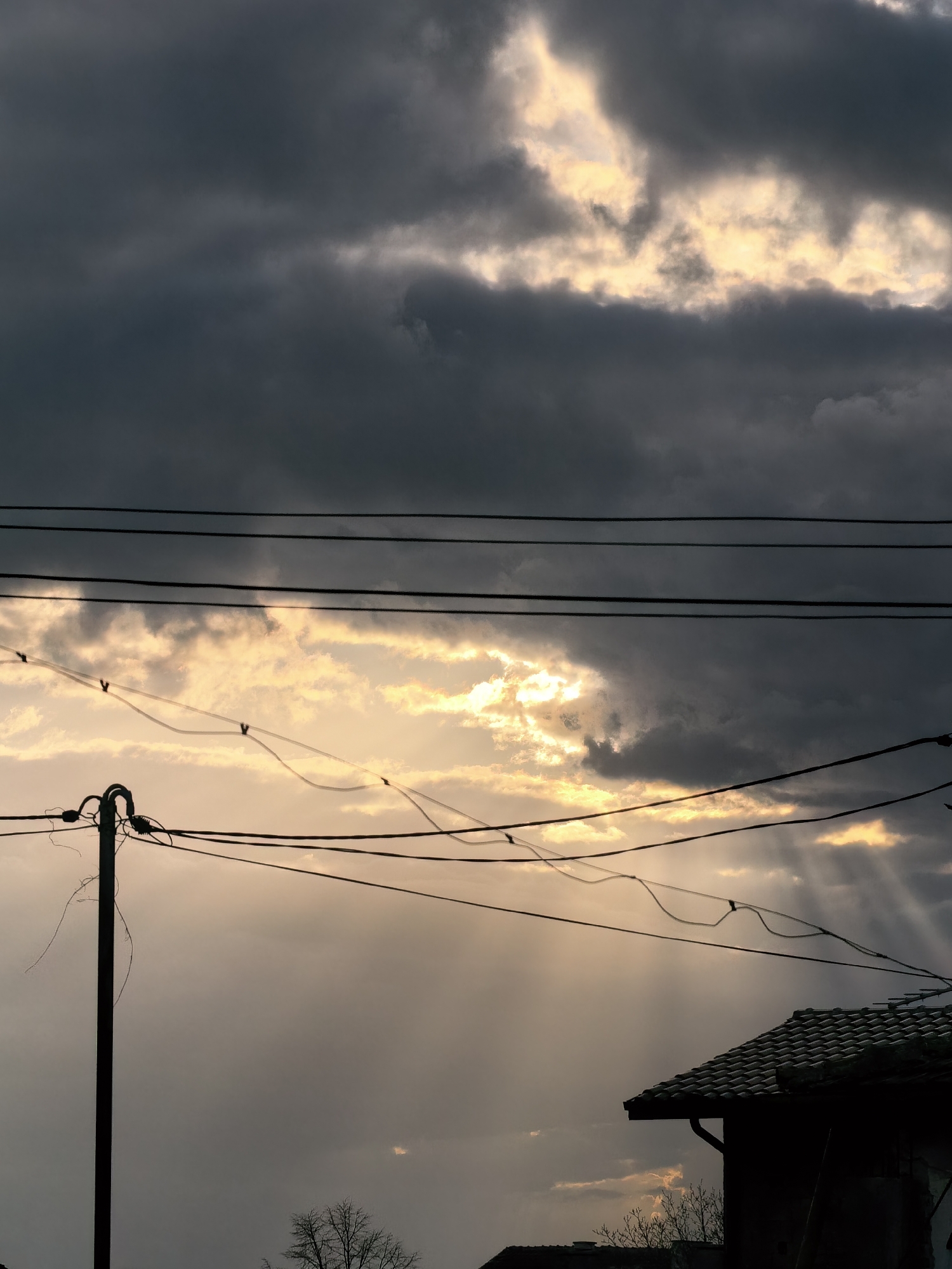 The image depicts a dramatic sky with dark, heavy clouds interspersed with patches of golden light breaking through. Sunlight streams down from the gaps in the clouds, creating a ray effect. In the foreground, there are several utility lines and a utility pole, casting a silhouette. The edge of a building with a tiled roof is visible in the lower right corner. In the distance, the bare branches of a tree can be seen. The location is urban but indistinct.