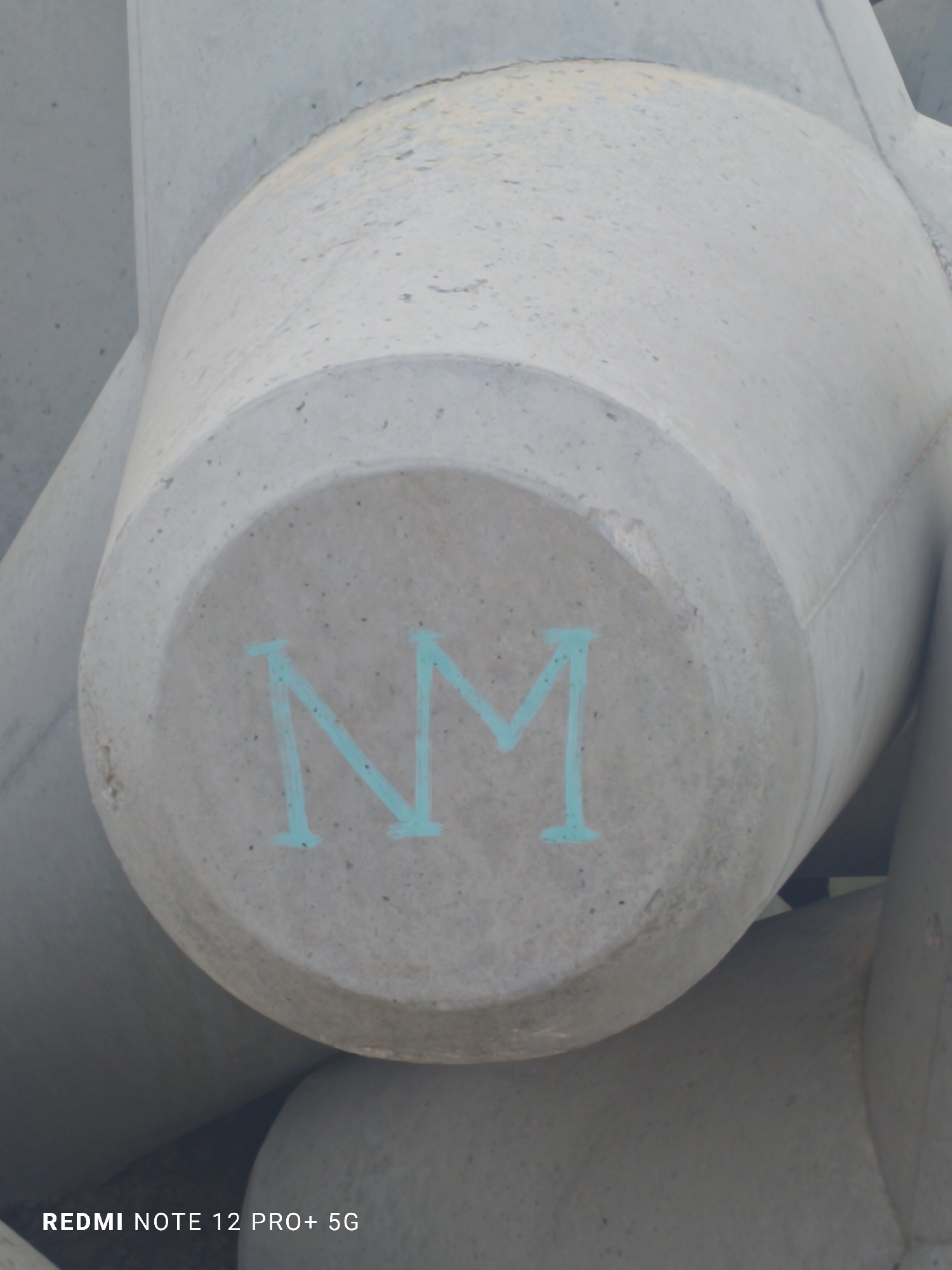The image captures a close-up view of a large, light grey concrete breakwater unit, specifically a type of tetrapod or similar pre-cast concrete block designed for coastal protection. The main subject is prominently featured, with its circular end facing the viewer. On this circular surface, the capital letters "NM" are stenciled or painted in a distinctive light blue or turquoise color.
In the background, other similar concrete structures are visible, appearing to interlock with the main block, suggesting a larger formation like a breakwater or sea defense. The overall scene is outdoors, consistent with the use of such heavy-duty concrete units in a maritime environment. The lighting indicates it is daytime, with diffused light suggesting either an overcast sky or indirect sunlight, as there are no harsh shadows. The concrete surfaces exhibit a textured, somewhat weathered appearance.
There are no people or explicit activities depicted in the frame. The visible text includes the painted "NM" on the concrete block and a digital watermark at the bottom left corner that reads "REDMI NOTE 12 PRO+ 5G," indicating the device used to take the photograph. Given the location context of Varna, Bulgaria, these structures are typically found along the Black Sea coast, serving to dissipate wave energy and prevent erosion.