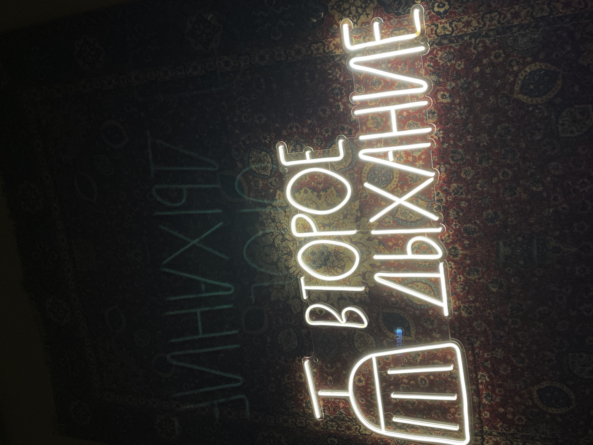 The image shows a neon sign displaying the Cyrillic text "ВТОРОЕ ДЫХАНИЕ" (Second Wind) with an abstract image below, photographed in what appears to be a room with a rug on the wall. The location is not discernible from the image.