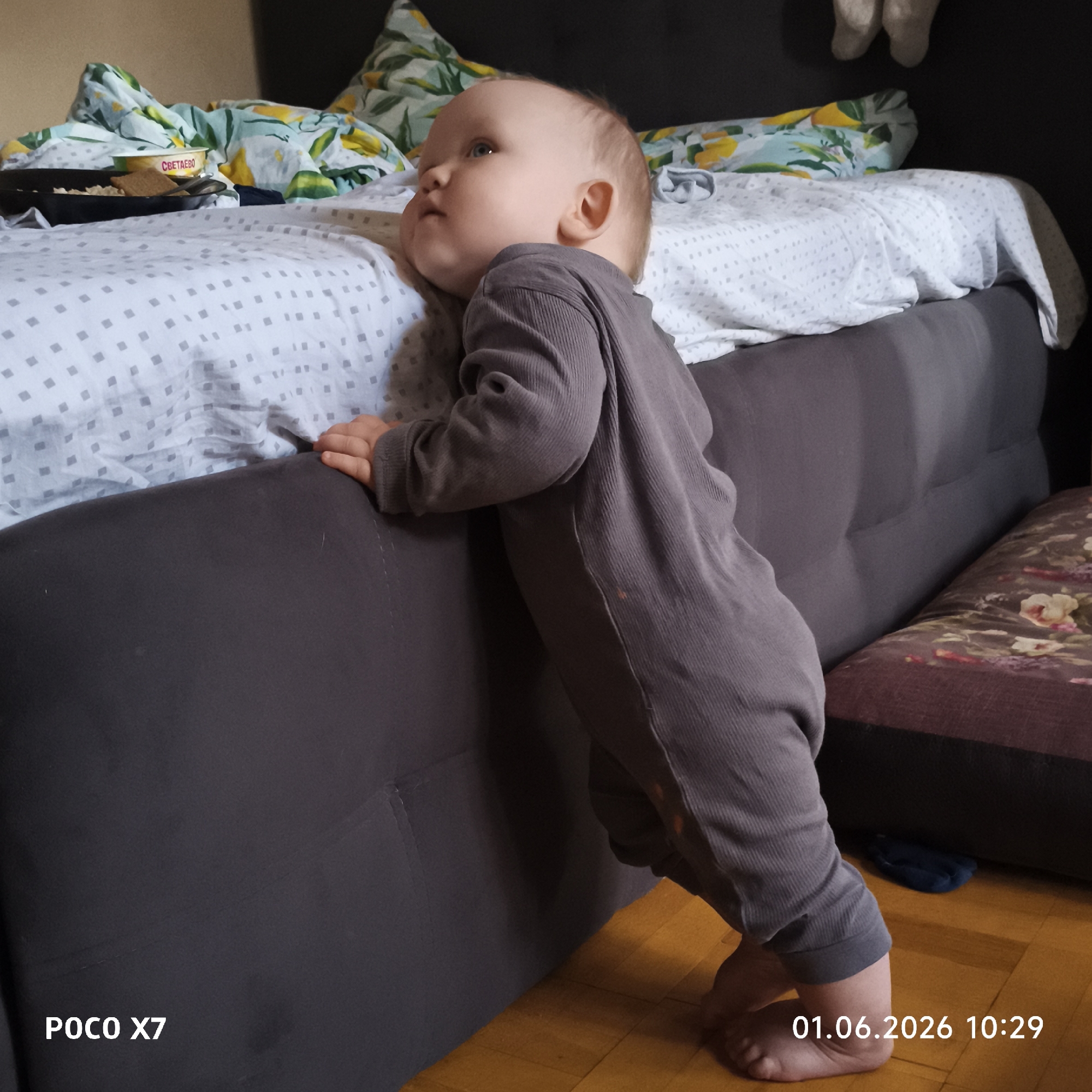 A young child, likely an infant, is standing and leaning against the side of a bed. The child is wearing a gray, corduroy-like jumpsuit. The bed has a gray headboard and a patterned sheet, with a pillow visible on top of the bed. A container of food is on the bed. The floor appears to be wooden. The bottom left corner of the image contains the text "POCO X7," and the bottom right corner reads "01.06.2026 10:29." The location is an interior setting.