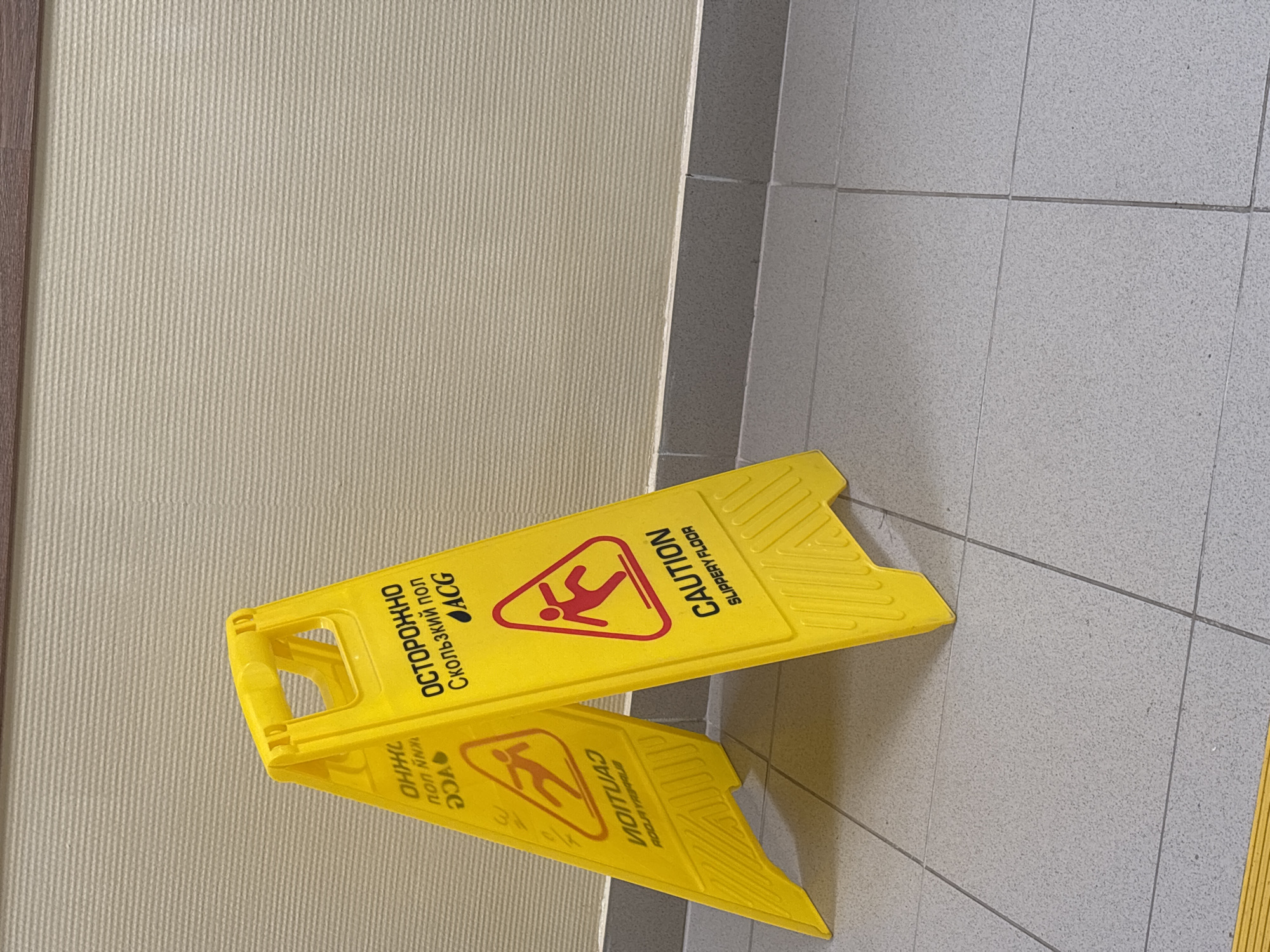 The image shows a yellow, A-frame "Caution Slippery Floor" sign placed on a tiled floor next to a wall with a textured, neutral-colored surface. The sign is labeled in both English and Russian ("ОСТОРОЖНО Скользкий пол"). The tile pattern on the floor suggests an indoor environment. The image does not provide enough information to determine the specific location in Chaikovsky, Russia.