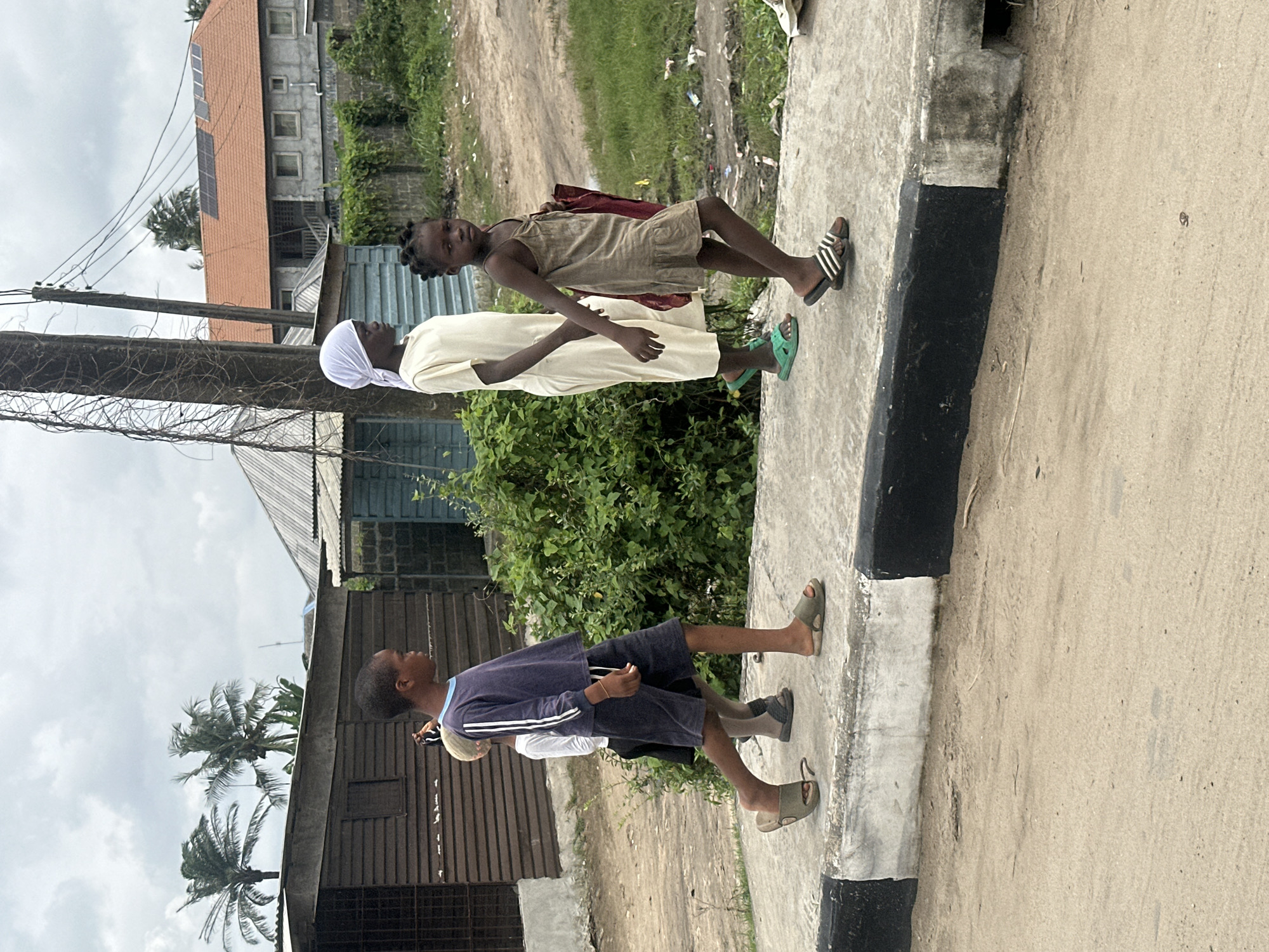 Four individuals are walking on a concrete sidewalk in front of a line of buildings. The first person on the left is a young boy wearing a purple long-sleeved shirt with white stripes and dark shorts, and brown sandals. He is walking to the left and appears to be carrying something on his shoulder. Behind him, a second young boy in a white t-shirt and dark shorts is also walking to the left. To the right of the first boy, a young girl in a light-colored, long-sleeved dress and a white headscarf is walking to the right, holding the hand of a third young girl. This third girl is wearing a light brown, sleeveless dress with ruffles at the hem and striped sandals. She is looking directly at the camera. The sidewalk is bordered by a black and white painted curb. Behind the sidewalk is a dirt path and overgrown green vegetation. In the background, there are several structures, including a wooden building on the far left, a blue building with a corrugated roof, and a larger building with an orange tiled roof and solar panels. Palm trees are visible on the left side of the image, and a utility pole with wires is present. The sky is overcast. This scene is located in Badagry, Nigeria.