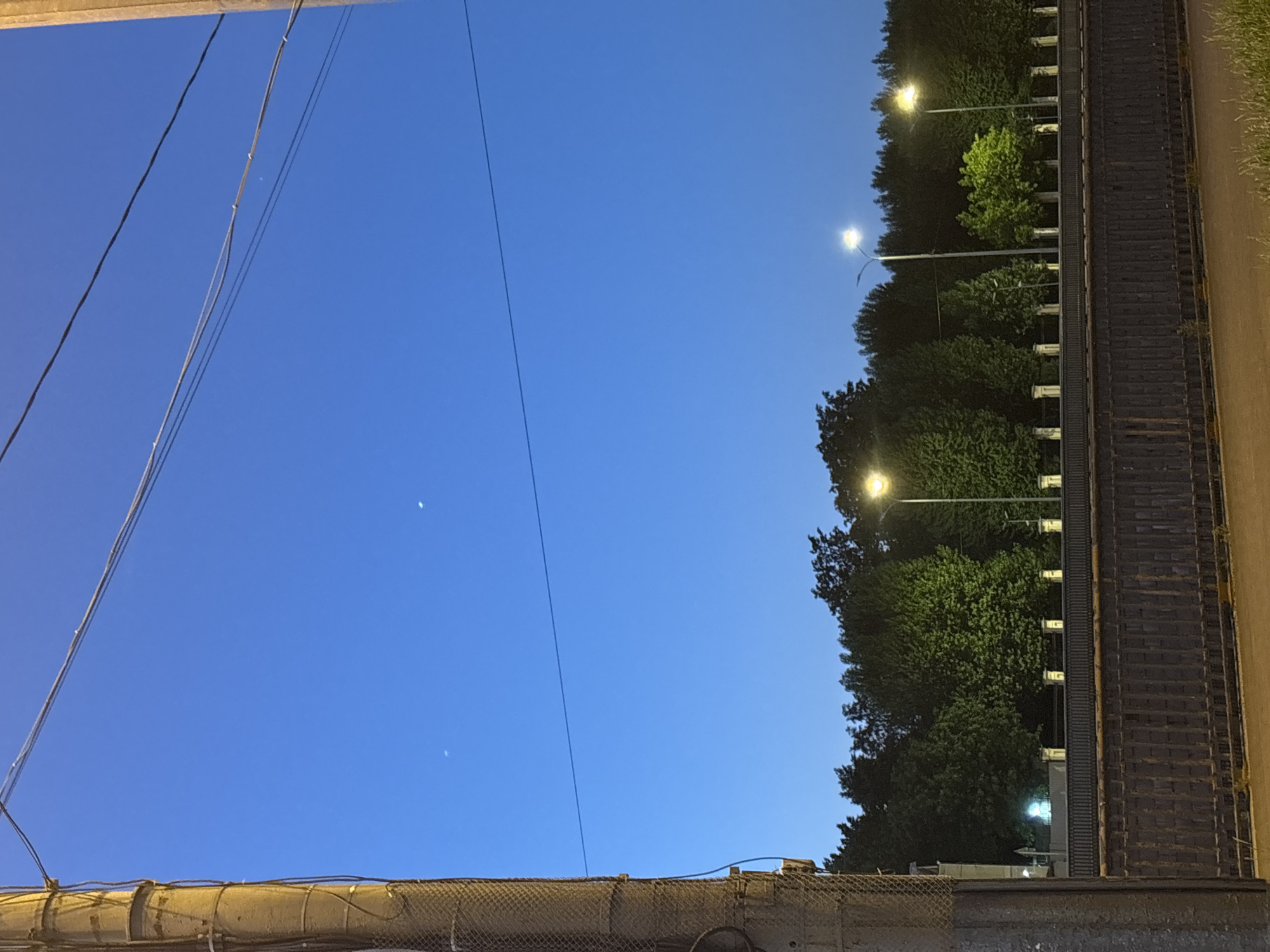 The image shows a street scene at dusk in Saint Petersburg, Russia. A tall, rough concrete utility pole with wires attached is on the left side of the frame. In the background, a line of trees is illuminated by streetlights. A dark, weathered fence or wall runs horizontally across the middle of the image, with decorative elements on top. The sky is a deep blue, with two faint celestial bodies visible.