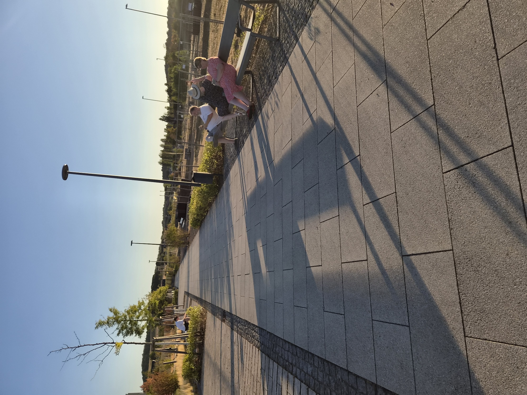 In Sanem, Luxembourg, on a clear, sunny day with a low-angle sun casting long shadows, a paved walkway runs diagonally from the foreground into the distance. The walkway is made of large, rectangular gray stone tiles with some smaller, dark gray cobblestones bordering it on the left. Along the left side of the walkway, there are young trees with thin trunks and some bushy green plants. On the right side of the walkway, a long, dark-colored park bench is occupied by three people. A man wearing a white t-shirt and shorts sits on the left end of the bench. Next to him, a woman in a dark dress and a straw hat is seated, holding up a smartphone as if taking a photo. On the far right of the bench, a woman in a pink dress is sitting. The background consists of a grassy area, more trees, and some distant structures, with a clear blue sky above. Several lampposts are visible along the walkway in the mid-ground.