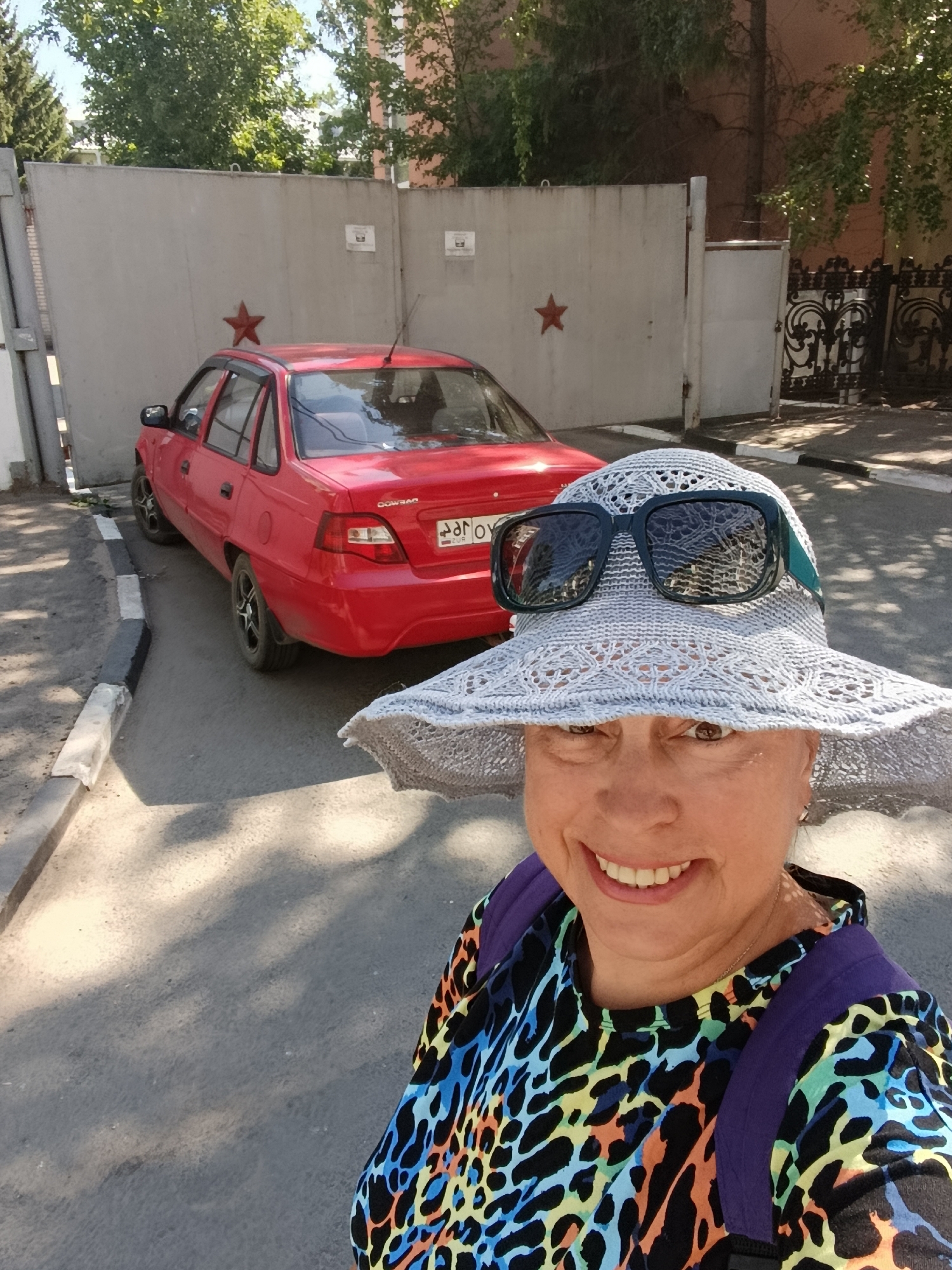 A woman wearing a wide-brimmed hat and sunglasses takes a selfie in front of a red car parked on a street in Saratov, Russia. The car is a Daewoo Nexia, and it is parked next to a concrete wall decorated with two red stars. In the background, there is a black metal fence and trees. The woman is smiling and wearing a colorful leopard-print shirt. She is also wearing a purple backpack. The sun is shining and casting shadows on the ground.