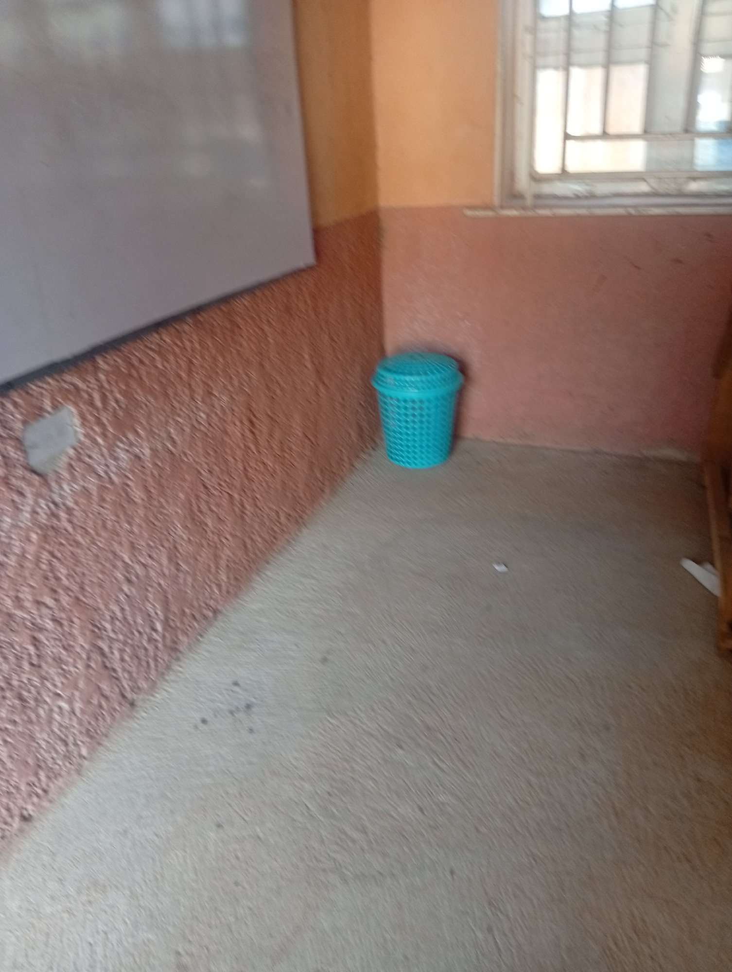The image depicts an indoor scene in Jalingo, Nigeria, likely a classroom or a room within a building. The walls are painted in two distinct colors: a peachy orange on the upper portion and a muted reddish-brown with a textured finish on the lower portion. To the left of the frame, a portion of a whiteboard or a similar flat, pale surface is visible, suggesting an educational or meeting setting.
In the center of the image, a bright turquoise plastic waste bin with a perforated design is placed on the floor. The floor itself is a light, neutral color with a slightly textured appearance, possibly linoleum or a similar flooring material. There are a few small dark specks on the floor, indicating minor dirt or debris.
To the right, a window with a metal security grate is visible, offering a glimpse of the outside. The light filtering through the window suggests it is daytime, but the weather conditions are not discernible. The overall impression is one of a functional, somewhat bare, interior space. No people or specific activities are evident in the frame, focusing instead on the static elements of the room. The image appears to be taken with a mobile phone camera, given the slightly tilted angle and the focus on everyday objects and environments.
