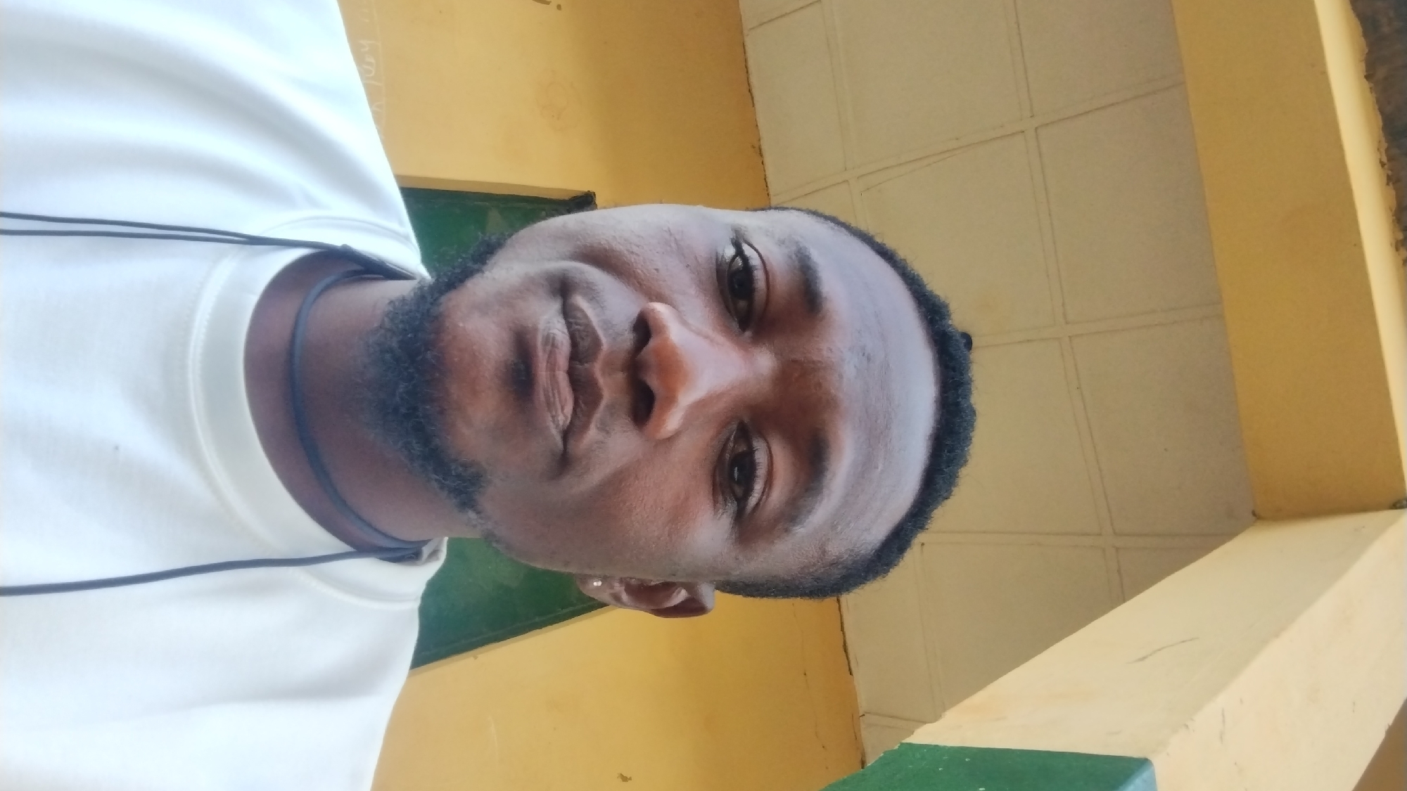 A close-up, head-and-shoulders portrait of a young Black man is captured from a slightly low angle. He has dark skin, short dark hair, and a neatly trimmed goatee and mustache. His eyes are dark, and his expression is neutral. He is wearing a white crew-neck sweatshirt and a thin black cord necklace. The background features a yellowish, textured wall and a yellow overhang with a white tiled ceiling, suggestive of an outdoor or semi-outdoor architectural setting. A hint of green can be seen to the right of the frame. This image appears to have been taken in Jos, Nigeria, though no specific landmarks are identifiable.