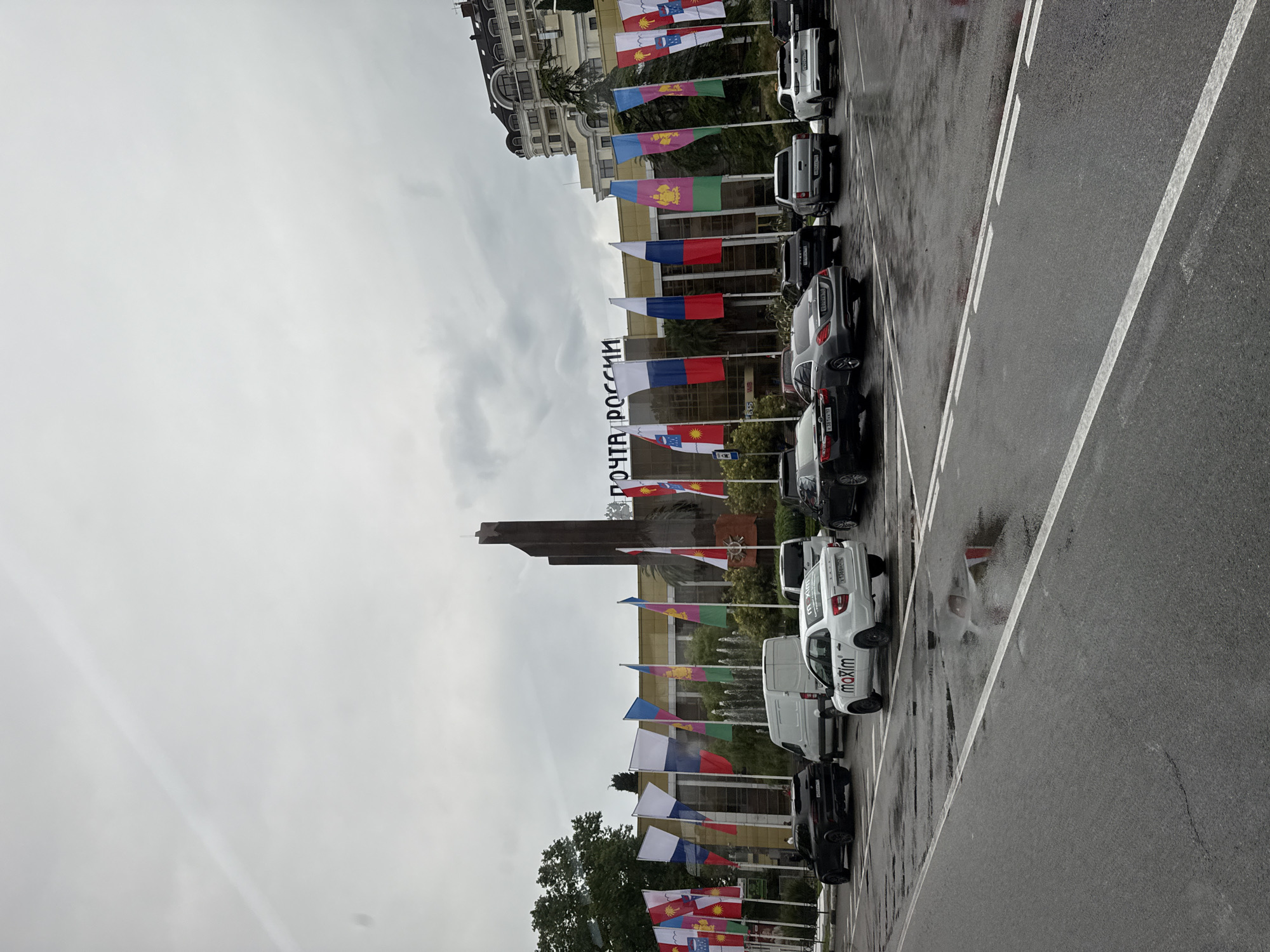 The image depicts the "Posta Rossii" building in Sochi, Russia, on a cloudy day. The building, which appears to be a post office, features a prominent sign with the words "ПОЧТА РОССИИ" (Posta Rossii) in large letters. In front of the building, a line of flags representing various Russian regions are displayed. Several cars are parked in the foreground and midground, including a white car with "maxim" branding, suggesting a taxi service. The ground in front of the cars is wet and reflective, indicating recent rain. The overall scene is that of a public space with a mix of official buildings and everyday transportation.